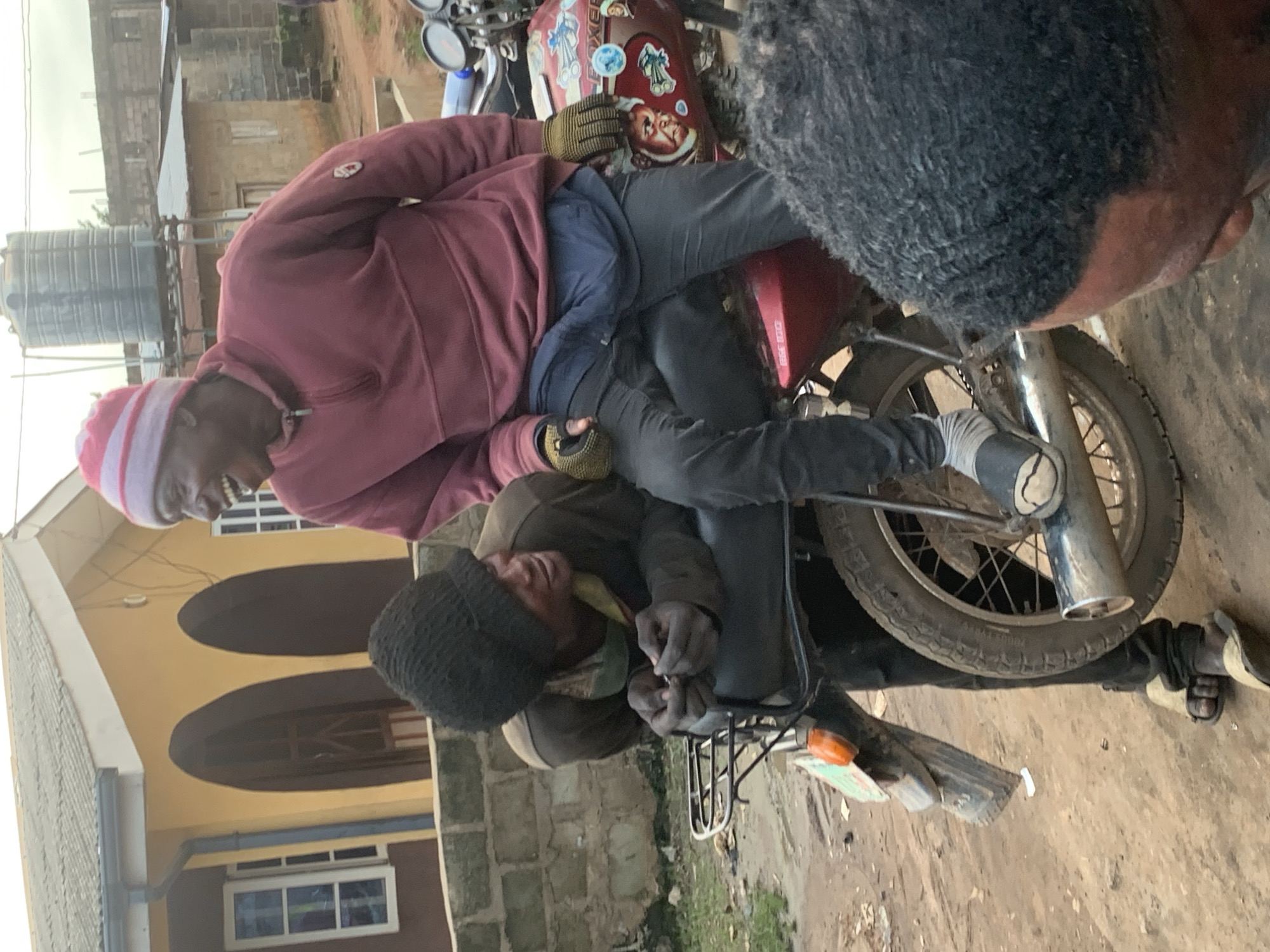 The image depicts an outdoor scene in the city of Shagamu, Nigeria, with a focus on two men positioned around a red motorcycle. One man, wearing a maroon sweatshirt and a striped beanie, is seated on the motorcycle and appears to be laughing, looking to his left. He is also wearing dark pants, grey socks, and what appear to be black slip-on shoes, with yellow gloves on his hands. The second man, in a dark beanie and a brown jacket over a light-colored shirt, is leaning against the back of the motorcycle, resting his forearms on the seat. His hands are clasped in front of him, and he is looking to his right. He is wearing dark pants and sandals. The motorcycle itself is red with various stickers on its side, including one that reads "BOXER." The background shows a yellow building with arched doorways and windows, and a large water tank on a metal structure. The ground is a dirt or gravel surface. Part of a third person's head and hair, viewed from behind, is visible in the foreground on the right side of the image.