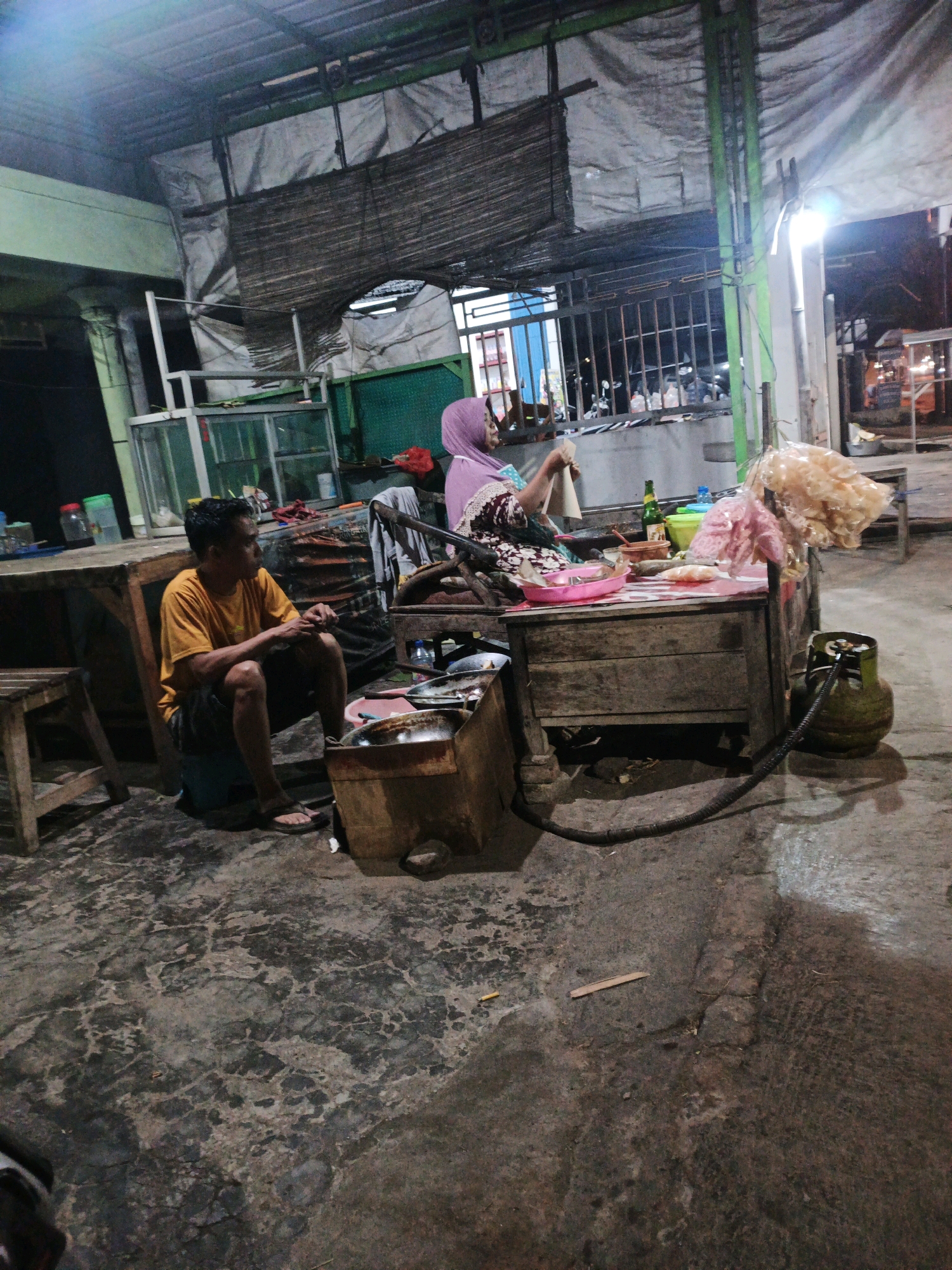 In Kintelan, Indonesia, an outdoor scene depicts a woman in a pink hijab and a patterned dress sitting behind a wooden table laden with food items and drinks. She is actively preparing or serving something. Beside her, a man in an orange t-shirt and dark shorts is seated on a blue stool, looking away from the table. The environment appears to be a food stall or an open-air eatery, with a corrugated metal roof overhead supported by green beams. A gas cylinder is visible next to the table, connected by a hose. The ground is a rough, concrete surface. The lighting suggests it is nighttime or dusk.
