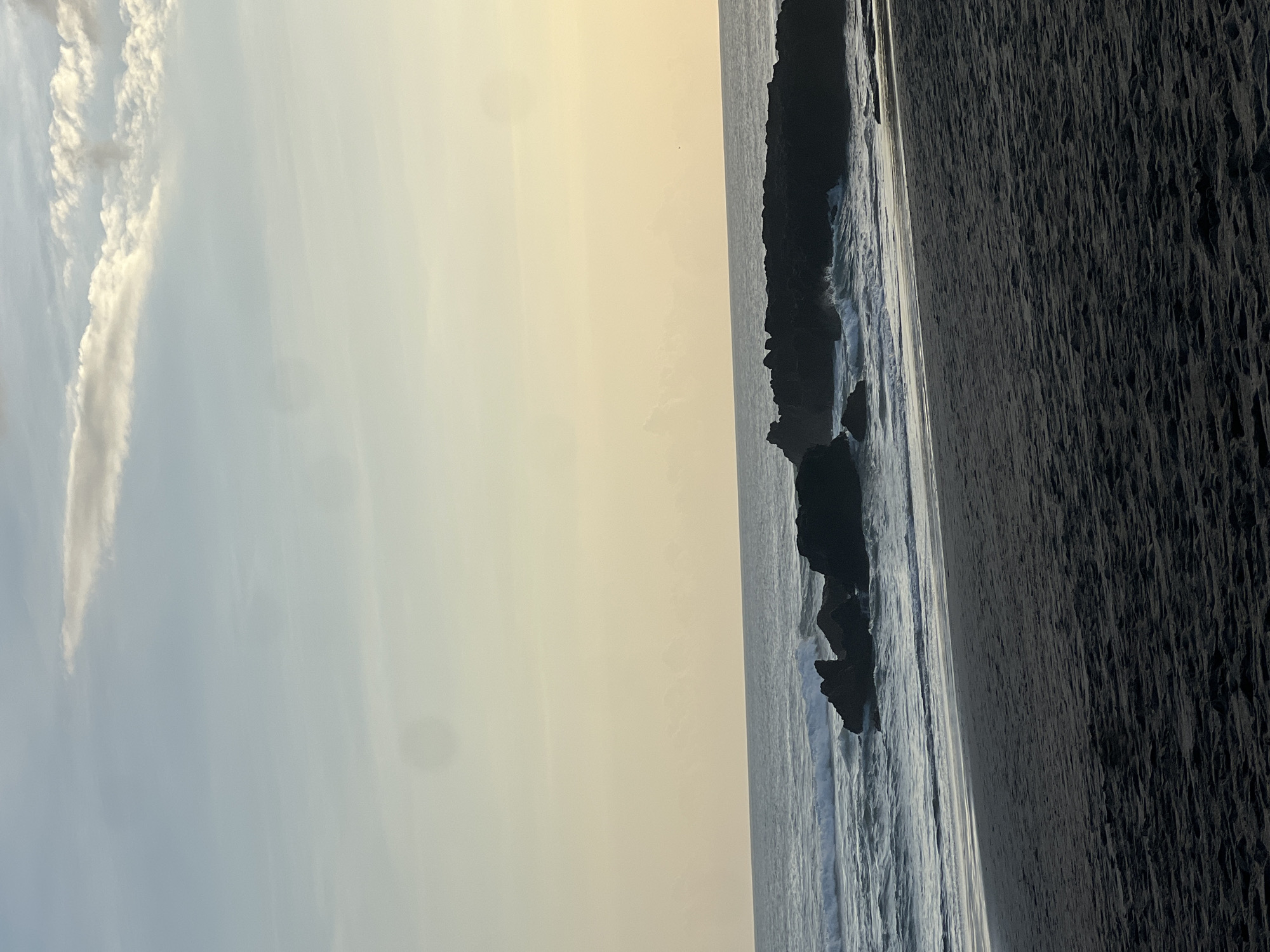 This is a photograph of a beach in North Kuta, Indonesia, at dusk. The sky is a soft gradient of blue and yellow, with thin, wispy clouds. The ocean is calm, with gentle waves rolling towards the shore. Several dark, rocky outcrops are visible in the water, with waves breaking around them. The beach itself is covered in dark sand. The image is taken from a low angle, looking out towards the sea. There are no people or animals visible in the frame.
