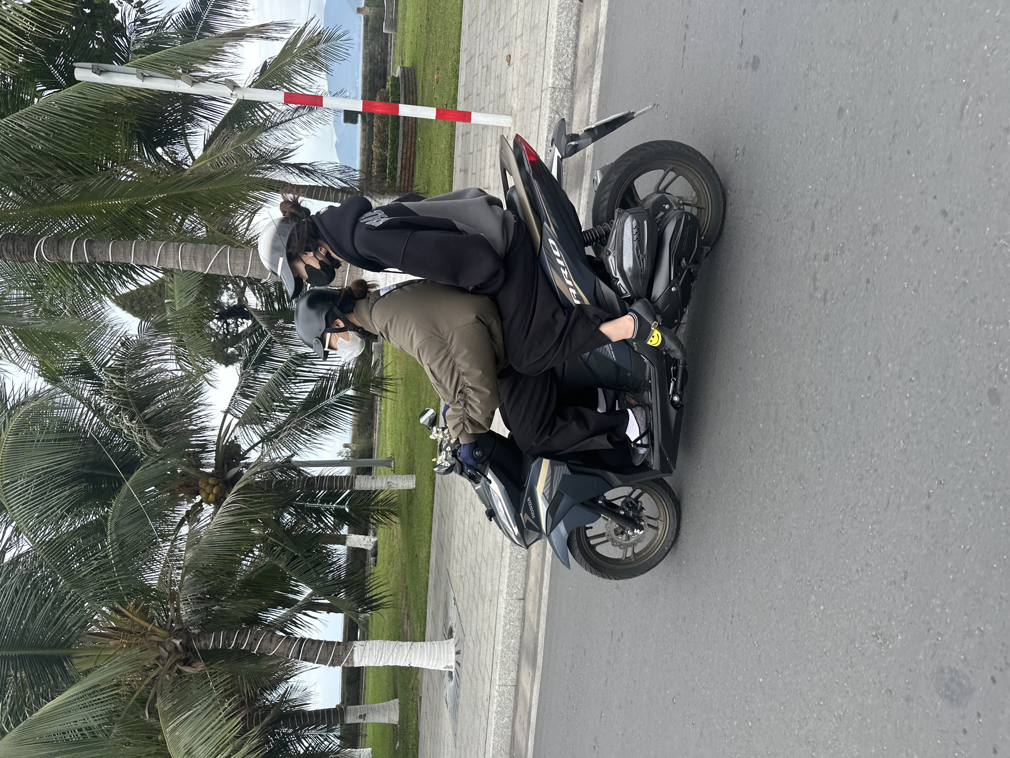 Two people on a black motorcycle are on a road next to palm trees and a walkway. Both are wearing helmets and face masks. The motorcycle has a luggage compartment on the back. In the background are a red and white striped barrier and greenery. The location is suggestive of Nha Trang Ward, Vietnam, based on the presence of palm trees and the red and white barrier.