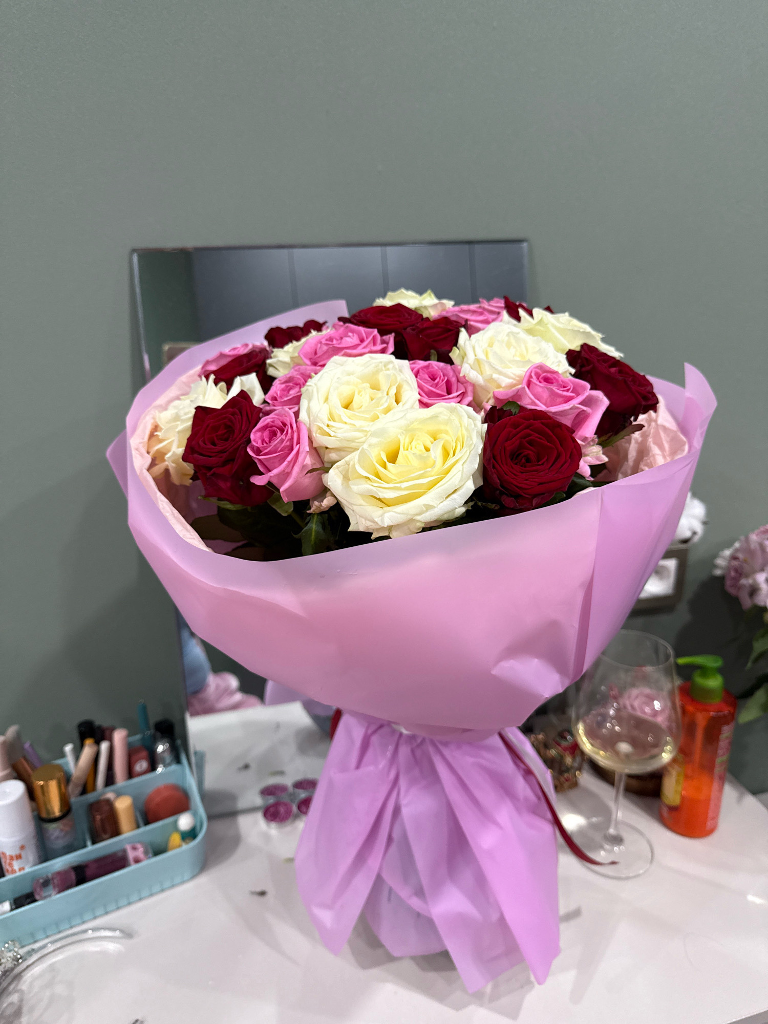 A bouquet of roses in various shades of red, pink, and cream is presented in a light pink paper wrap, tied with a red ribbon. The bouquet is positioned on a white surface, likely a table or dresser, which also holds a clear wine glass containing a pale yellow liquid and a bright orange bottle with a green dispenser. To the left of the bouquet, a light blue organizer is filled with makeup items, including lipsticks, brushes, and small bottles. A portion of a mirror is visible in the background, reflecting the bouquet and the wall behind it, which is painted a muted green. The scene appears to be indoors, possibly a bedroom or dressing area. No people are present in the image. This scene is not in Cheboksary, Russia.