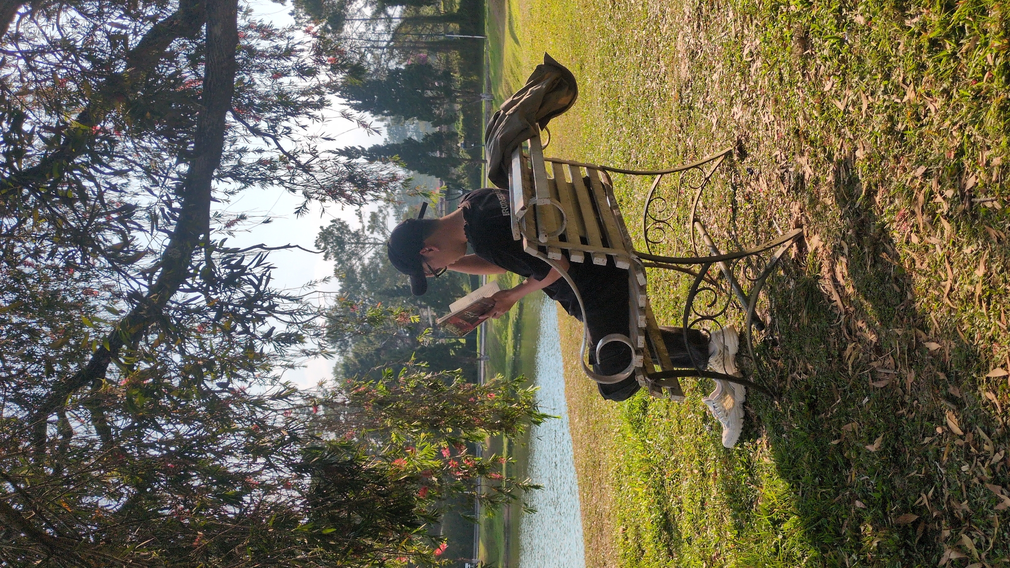 An individual wearing a black baseball cap, glasses, and a black shirt sits on a park bench beside a lake, reading a book. A jacket is draped over the back of the bench. The scene appears to be outdoors, with the bench situated on a grassy area, partially covered with fallen leaves. The location is Da Lat, Vietnam, as suggested by the environment. Foliage and trees surround the area, some obscuring the lake. The person is white, wearing white sneakers.