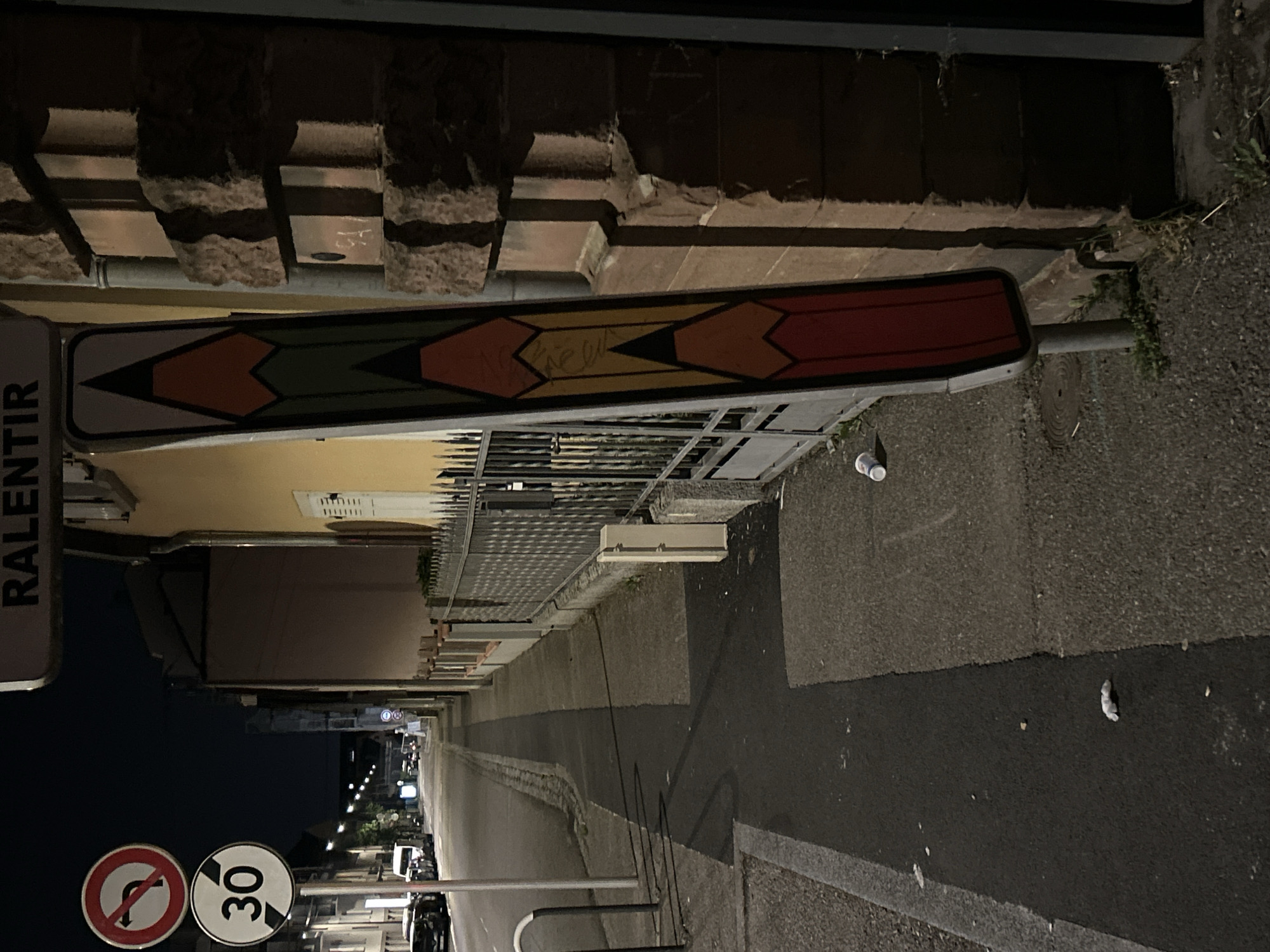 The image is a street scene at night in Strasbourg, France. A speed limit sign of 30 is visible, along with a "no u-turn" sign and a "ralentir" sign. A long, horizontal sign with a series of colored arrows (white, orange, green, yellow, red) points towards the street's center. The street is lined with buildings and a metal fence. The pavement is visible with some litter. Street lights illuminate the scene.