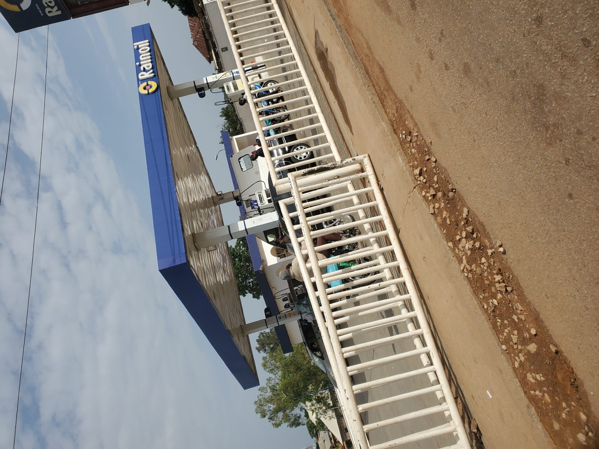 This is a medium-shot, angled, outdoor photograph of a Rainoil gas station in Bukuru, Nigeria. The gas station has a blue canopy with the Rainoil logo visible. Several vehicles, including a car and at least two motorcycles, are present at the pumps. People are seen operating the pumps or seated on the motorcycles. The scene is viewed through a white metal fence that runs across the foreground. The ground is paved, and there is some debris and patchy vegetation visible. The sky is partly cloudy.
