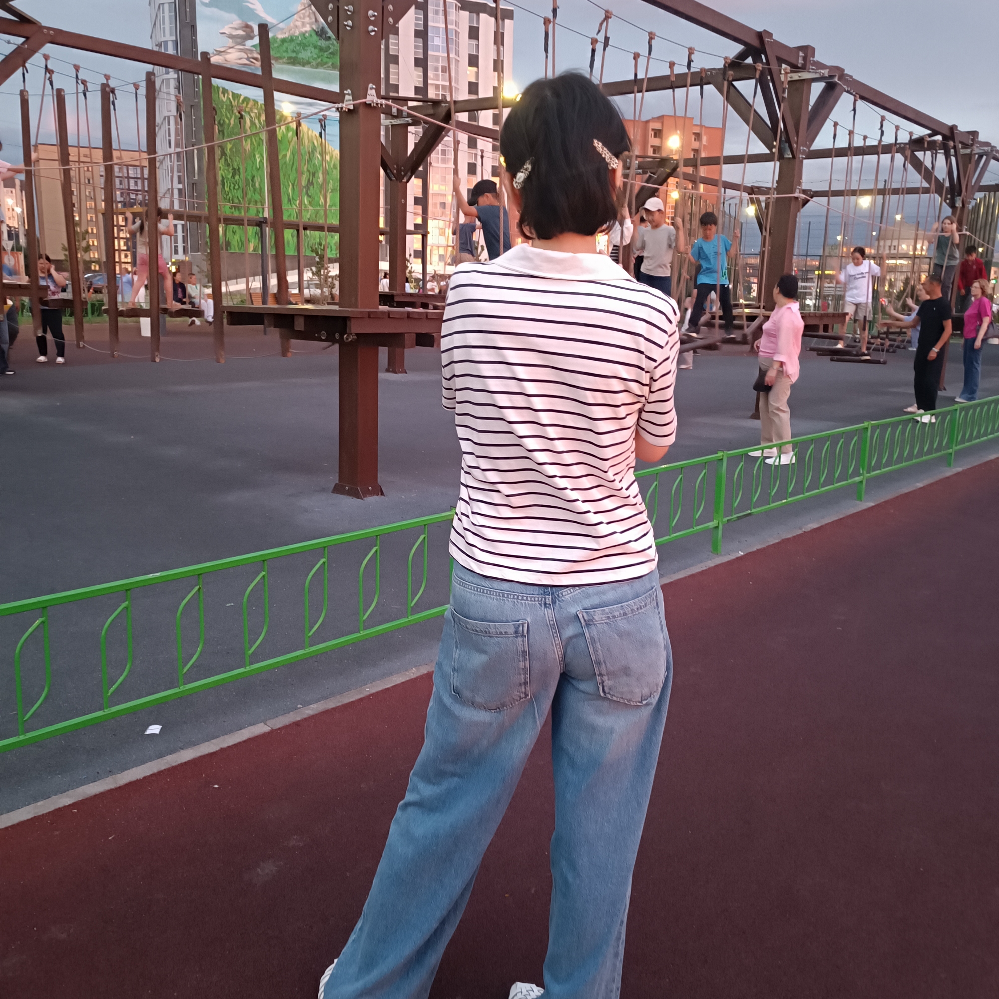 This image captures an outdoor adventure park, likely a ropes course, with several people, predominantly children, engaged in the activity. The main subject is a person viewed from behind, wearing a horizontally striped long-sleeved shirt and light blue jeans. They have short dark hair styled with two decorative clips.
The ropes course itself is constructed from dark wooden beams and supported by sturdy metal posts. Numerous ropes and suspended elements, such as wooden planks and what appear to be climbing nets, form the challenging pathways. Several children are visible on the course, navigating its various obstacles. One child in a light blue t-shirt and dark pants is seen in motion, while others in the background are also participating. Adults are present, some observing from the ground and others on the course.
The setting appears to be an urban park, with tall buildings visible in the background. The sky suggests it is late afternoon or early evening, with a soft, diffused light indicating possibly overcast conditions or the approaching twilight. The ground is a dark asphalt surface, with a bright green metal fence running along the edge of the play area. The overall atmosphere seems to be one of recreation and outdoor activity, with the focus on the adventurous nature of the ropes course. No text is visible in the image.