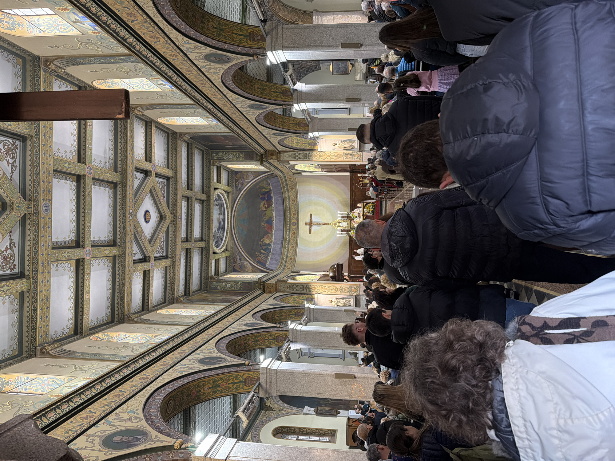 The image shows the interior of a church, filled with people attending a service. The church has a highly decorated ceiling with geometric patterns, arches, and religious artwork. Light streams in through the windows. A crucifix is visible at the altar. The location is likely a church in Plovdiv, Bulgaria.