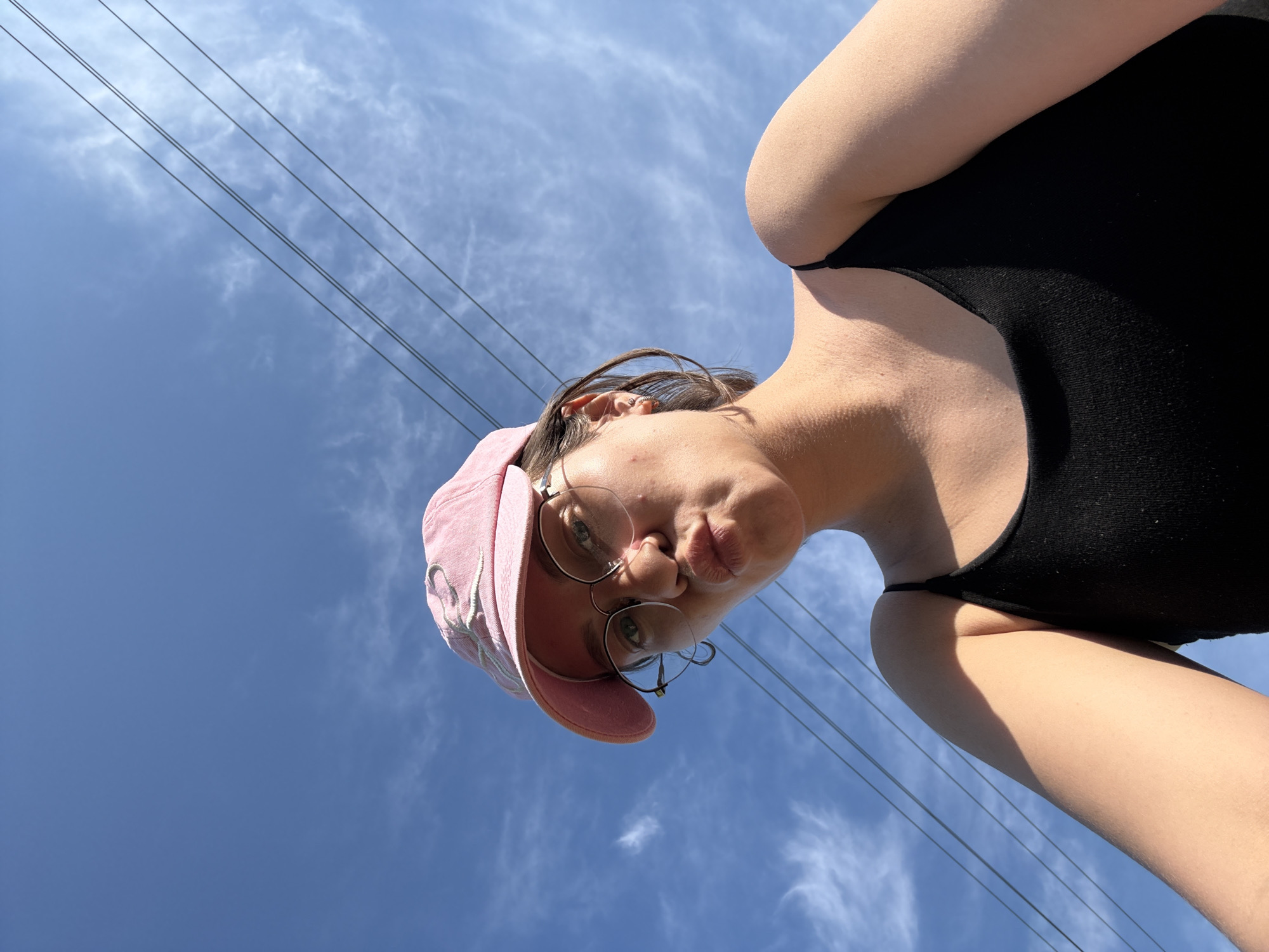 A low-angle, full-body shot from below the subject's chin upwards shows a young woman wearing a pink baseball cap and a black tank top, with a bright blue sky filled with wispy clouds and power lines in the background. She is looking upwards towards the camera with her lips pursed in a kiss. Her round glasses rest on her nose, and her short brown hair is visible beneath the cap. The scene appears to be outdoors, with natural sunlight illuminating the woman. The power lines stretch horizontally across the upper portion of the frame, suggesting an urban or suburban environment, possibly in the city of Dokshitsy, Belarus, given the user's prompt.
