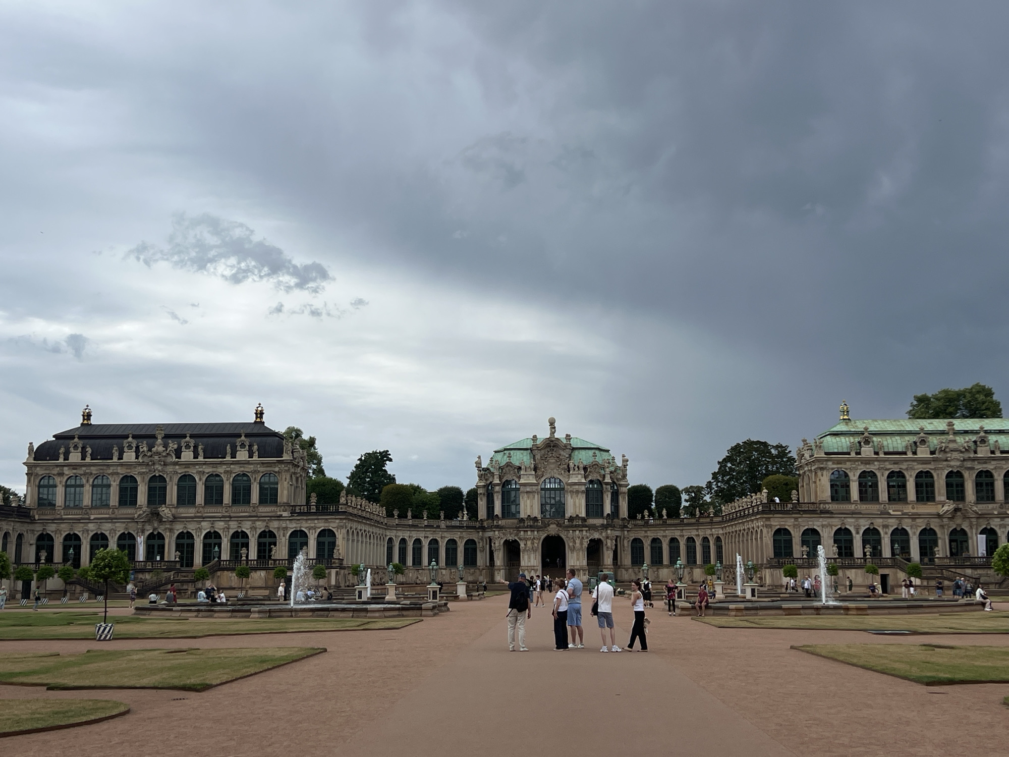 This image captures the Zwinger in Dresden, Germany, a baroque palace complex known for its gardens and museums. In the foreground, a group of people stands on a wide, reddish-brown path. The architecture of the Zwinger is visible in the background, characterized by its ornate facades, arcades, and green-domed roofs. Several fountains are active within the courtyard, with water spraying upwards. The sky is overcast with heavy grey clouds, suggesting impending rain or a storm. The overall scene depicts a public space with a mix of architectural grandeur and natural elements, under a dramatic sky.