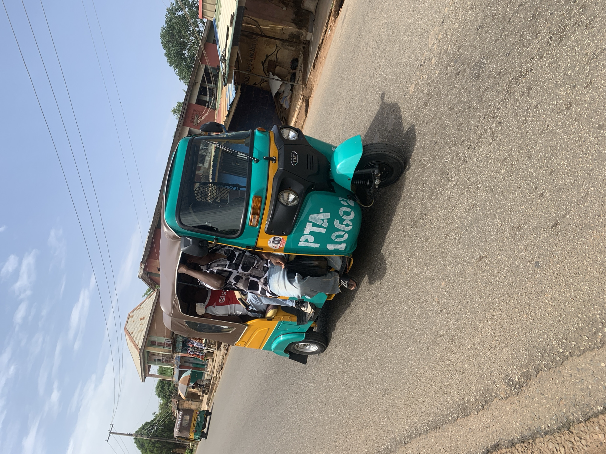 A motorized tricycle taxi, painted in a combination of yellow, brown, and teal, is carrying passengers on a paved road in Gura Top, Nigeria. The taxi is facing towards the top-right of the frame. On the side of the taxi are the letters "PTA-10602" in white paint. The driver, visible through the front window, is wearing a black and white patterned shirt. A passenger wearing a red and white shirt is also visible. The road occupies most of the frame, with buildings and trees visible in the background. Overhead electrical wires are also visible against a light blue sky.