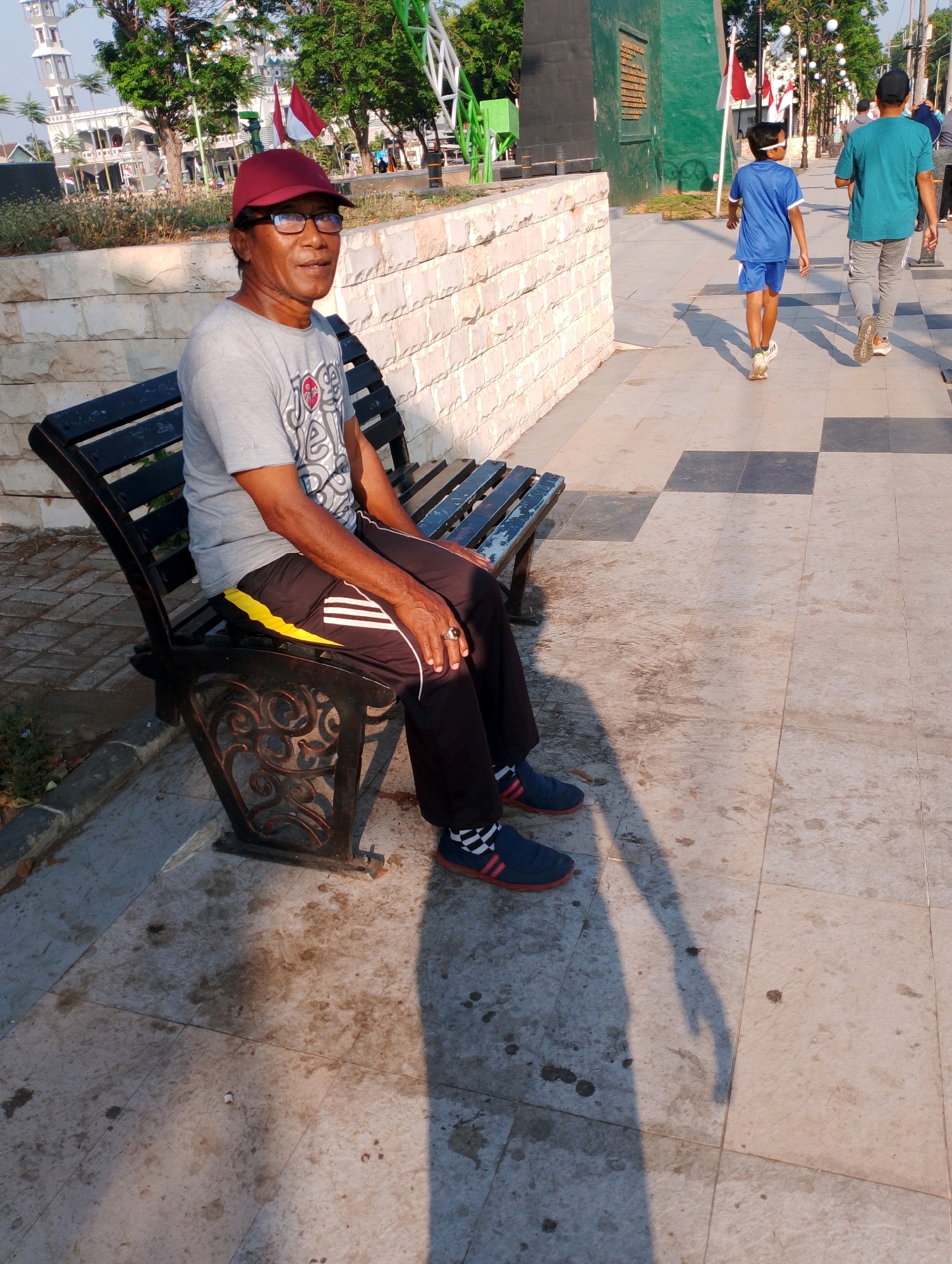 The image captures a man wearing a red cap, glasses, a gray t-shirt with a graphic print, black pants with yellow and white stripes, and blue athletic shoes with red trim and checkered socks, seated on a black metal park bench. He is positioned on the left side of the frame. In the background, two young boys in athletic wear are walking away from the camera on a paved walkway. The walkway features a checkerboard pattern of light and dark square tiles. Behind the boys and the walkway is a low, textured light-colored wall and further back, a building resembling a mosque with a minaret, trees, and flags. The scene appears to be an outdoor public area, possibly a park or promenade, with abundant sunlight casting long shadows. The location is identified as Probolinggo, Indonesia.