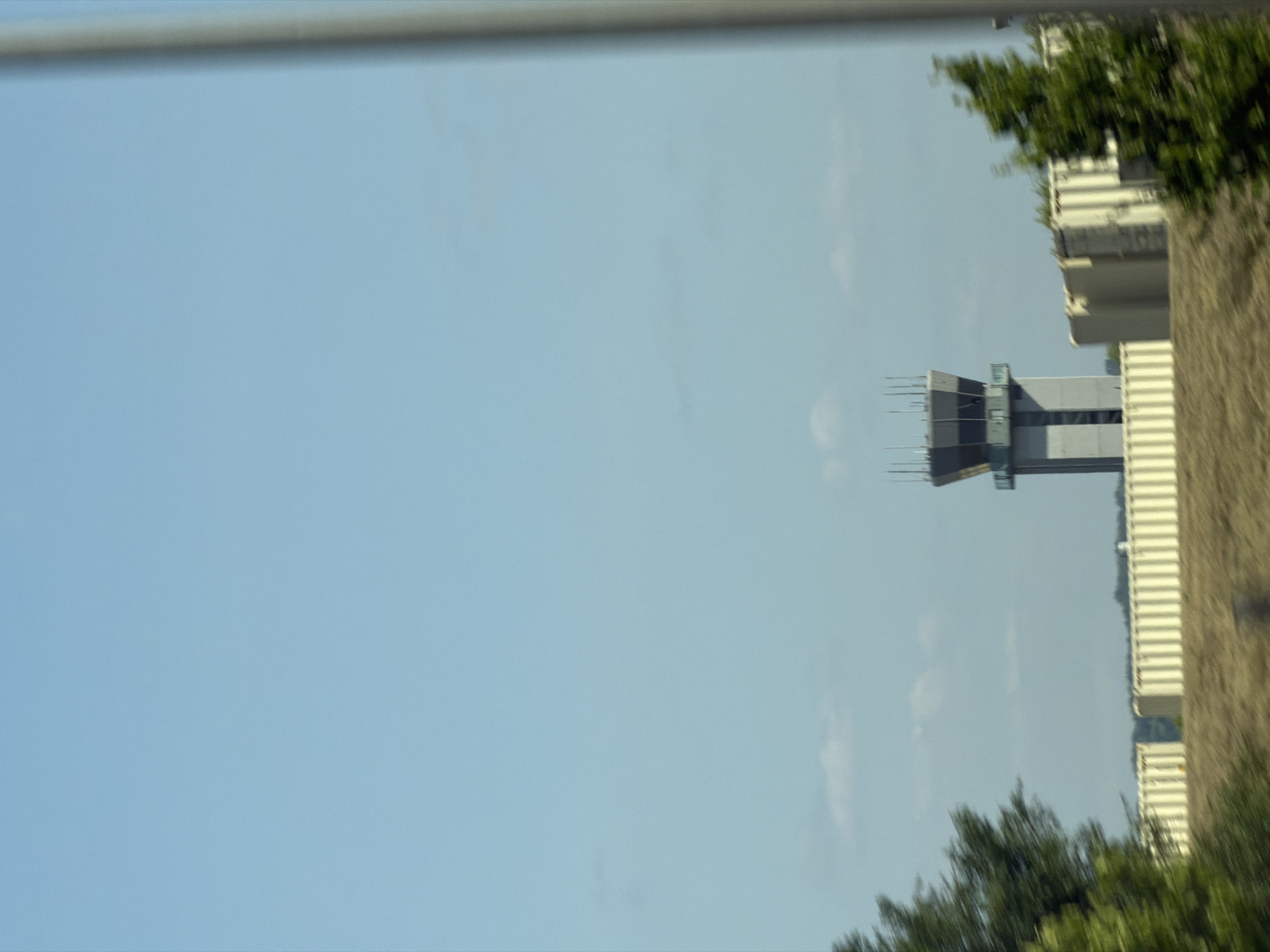The image shows an airport control tower in Saint Paul, United States of America. The control tower is tall and octagonal, with a glass-enclosed cab at the top. It is surrounded by several low-lying buildings, which appear to be hangars or administrative offices. The sky is clear and blue, with a few scattered clouds. Trees and vegetation are visible in the foreground, and a blurry metal pole is present on the right side of the image. The overall scene suggests an active airport environment on a clear day.