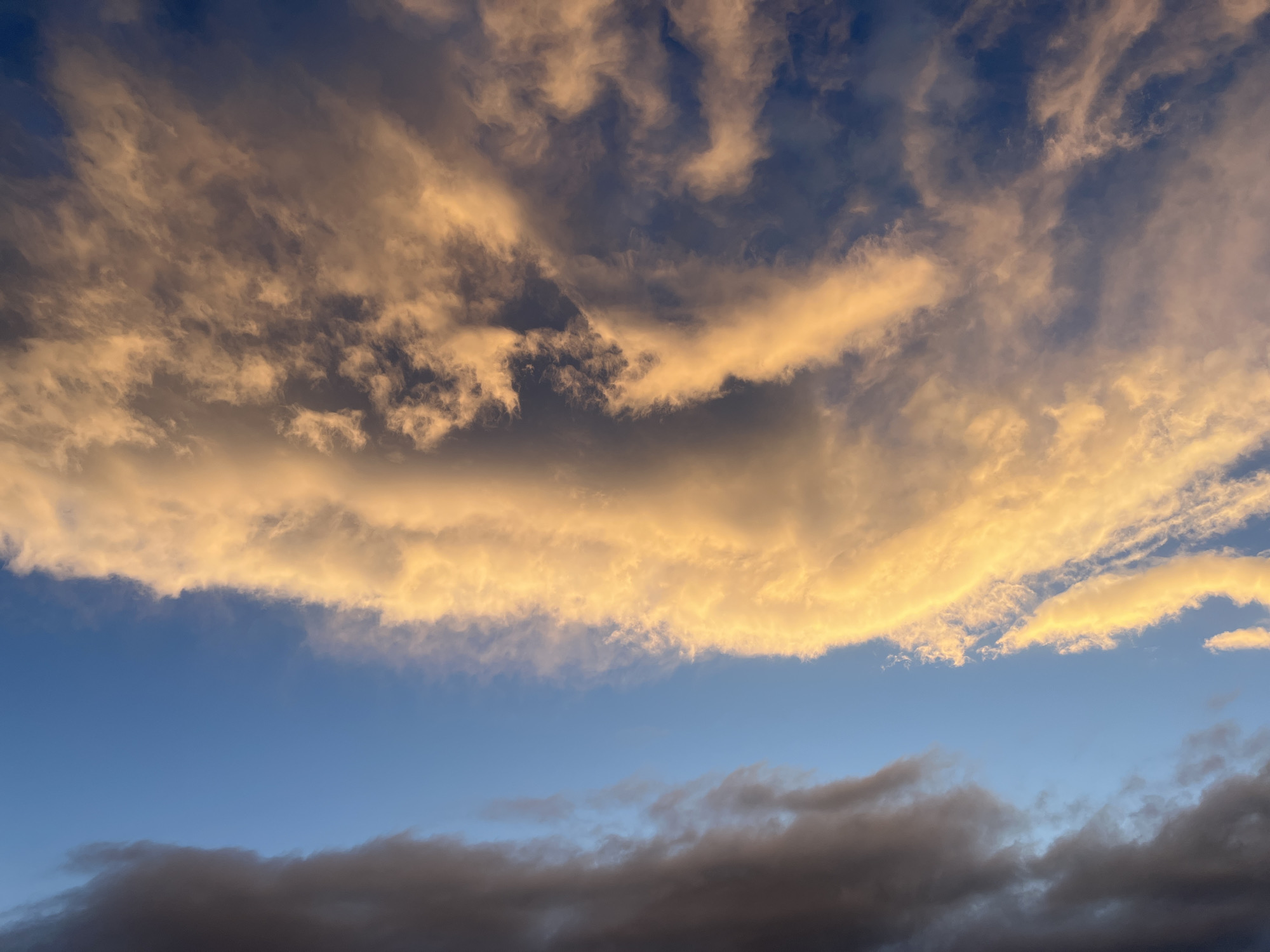 The image shows a sky in Aparan, Armenia. The upper half is dominated by clouds illuminated in hues of gold and tan. Below these clouds is a band of blue sky, followed by a lower layer of darker, grey clouds.