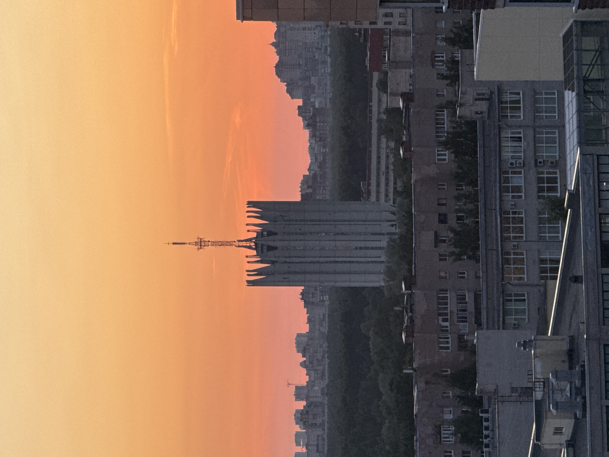 The image captures an urban landscape at either sunrise or sunset, bathed in a warm gradient of orange and yellow light. The sky, occupying the upper portion of the frame, transitions from a soft yellow at the top to a deeper orange-red closer to the horizon.
In the mid-ground, a distinctive tall, slender structure, resembling a communication or television tower, stands prominently with an antenna at its peak. Adjacent to it, a large, imposing building with a unique, angular architectural style, possibly Soviet-era, is visible. Further in the mid-ground and background, a sprawling cityscape unfolds, with numerous multi-story buildings forming a dense skyline, mostly silhouetted against the bright sky. A band of dark green, indicating trees or a park, runs horizontally through the mid-section, separating different urban elements.
In the foreground, on the right side of the image, several modern multi-story buildings are detailed. Their facades feature numerous windows, some reflecting the warm sky, and visible air conditioning units. The architectural style suggests an Eastern European or post-Soviet context, aligning with the given location hint of Russia.
The scene is devoid of people or discernible activity, presenting a serene and expansive view of the city as it transitions between day and night. No visible text is present in the image.