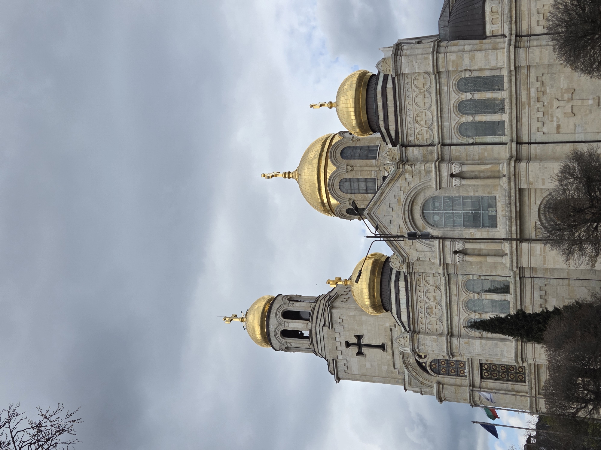 The image shows a low-angle view of the Dormition of the Mother of God Cathedral in Varna, Bulgaria. The cathedral is a large, ornate building with several golden domes topped with crosses. The facade is decorated with crosses, arched windows, and other architectural details. The sky is cloudy and overcast. In the foreground, there are bare tree branches and some flags.