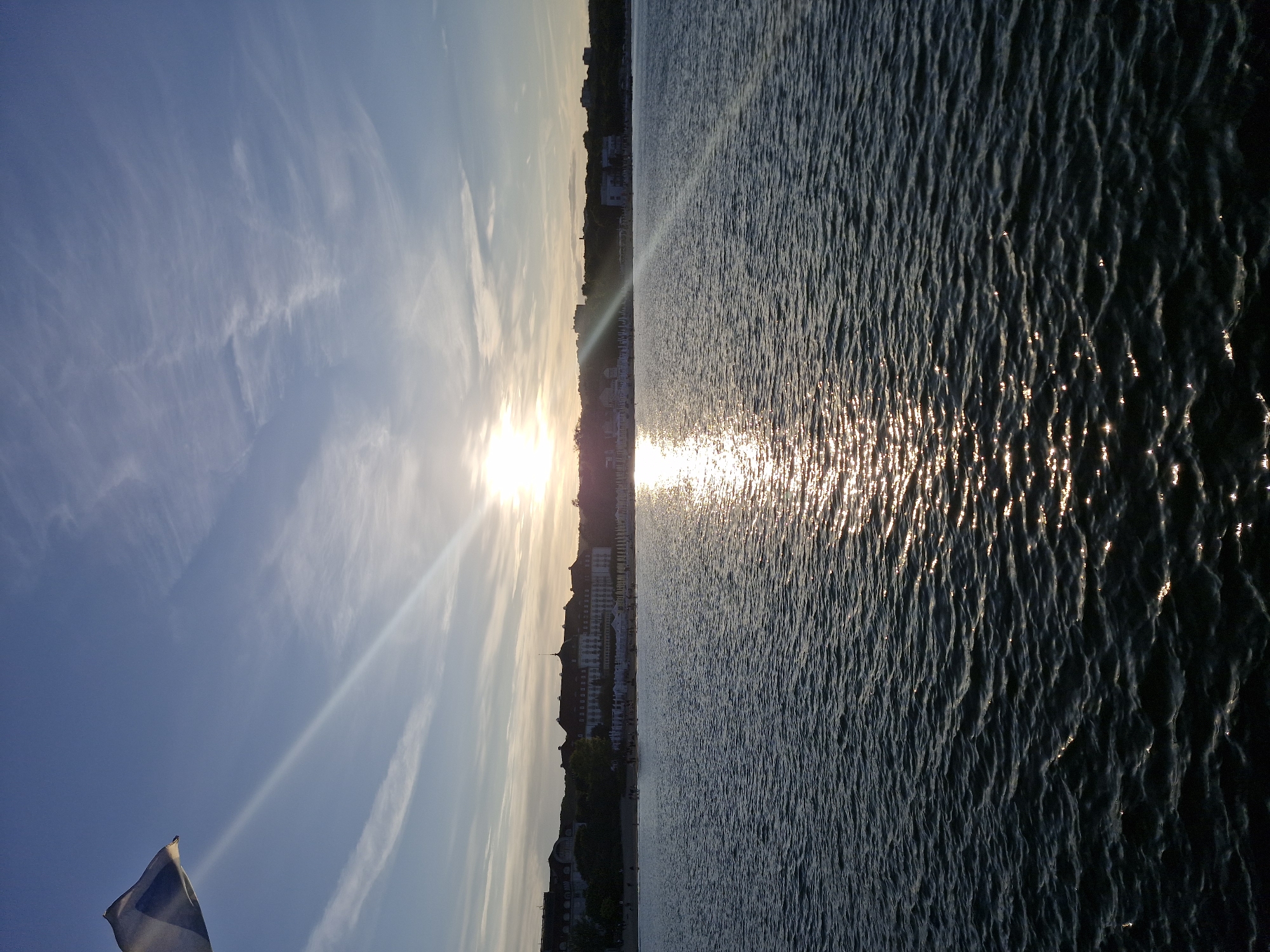The image depicts a seaside view of Sopot, Poland, at sunset. The sun, a bright orb, is visible near the horizon, casting a golden shimmer across the rippling water of the Baltic Sea. The light rays emanating from the sun are clearly defined, streaking across the sky. In the foreground, the dark blue water is textured with small waves reflecting the sunlight.
In the middle ground, a sandy beach stretches along the coastline, dotted with numerous white beach umbrellas. Beyond the beach, a line of buildings with red-tiled roofs forms the Sopot shoreline, suggesting a developed urban area. Some trees are visible behind the buildings, and a few scattered figures can be discerned on the beach, indicating human presence.
In the upper left corner of the image, a white and blue flag is partially visible, waving in the wind, likely attached to a boat or a pier. The sky is a gradient of blue, transitioning to lighter shades near the horizon, with wispy white clouds present. The overall scene captures a serene and picturesque sunset over the Sopot coastline.