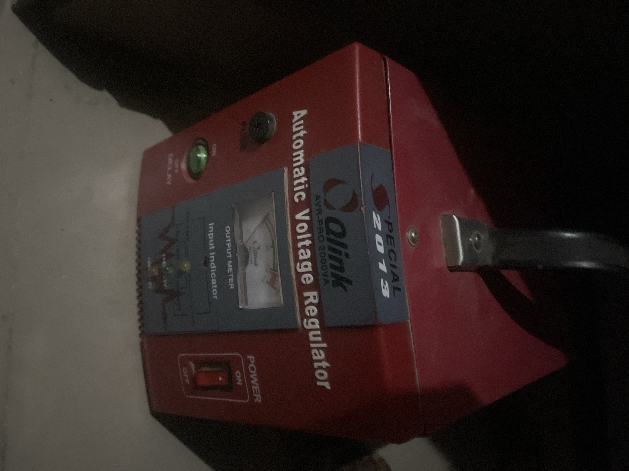 The image shows a red voltage regulator, a Qlink AVR-PRO 2000VA, with a handle on top. It is labeled "SPECIAL 2013". The device has an output meter, an input indicator, and a power switch. There are also buttons labeled "FUSE", "ON", "OFF DELAY". The device appears to be in use, as a green light is illuminated. The background is dark, suggesting it is nighttime or indoors with minimal lighting. The location is identified as Jalingo, Nigeria.