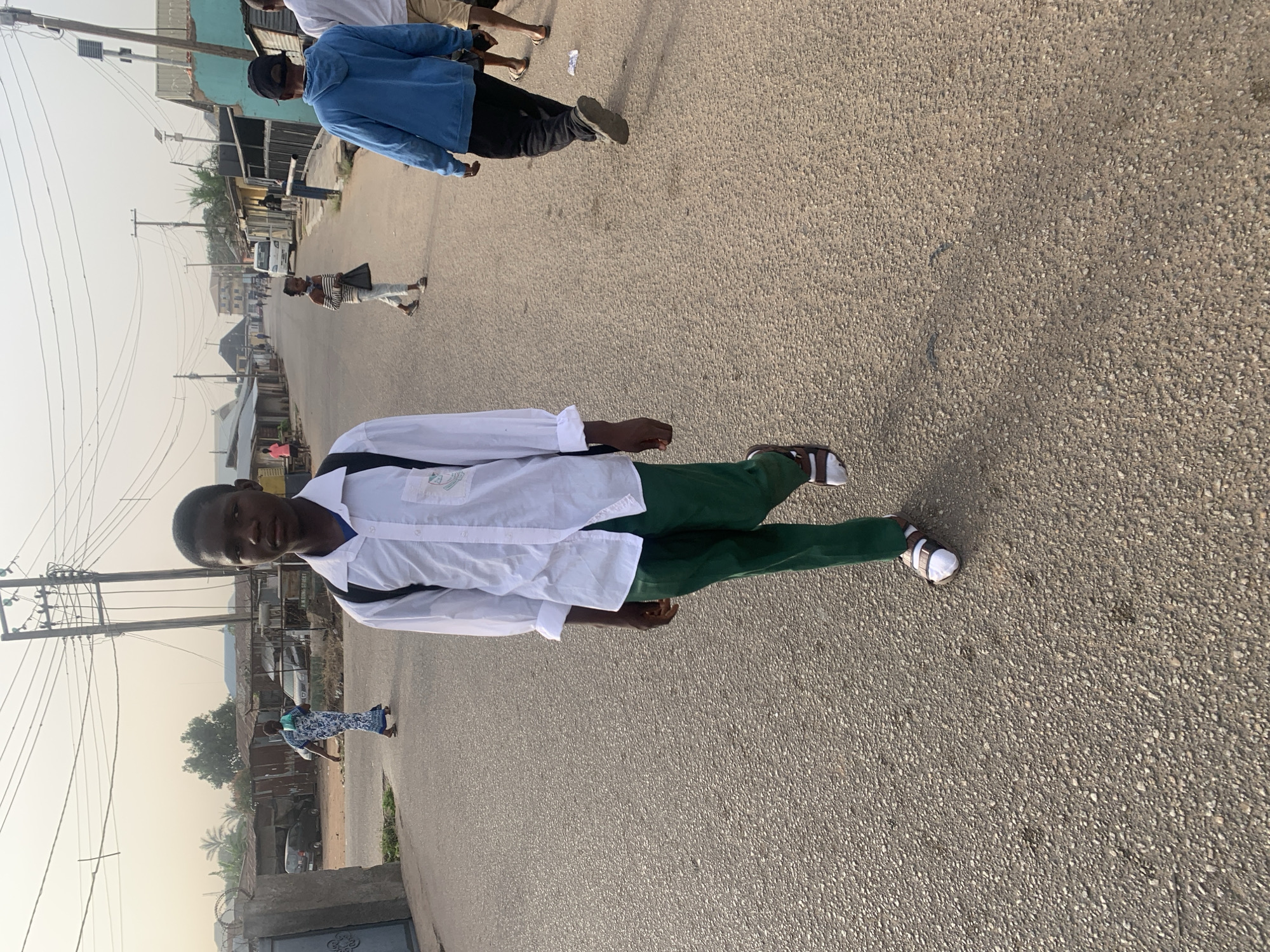 The photograph shows a young man dressed in a white shirt and green pants, standing on an asphalt road. He's wearing sandals. He is facing the camera with a neutral expression. In the background, there are buildings, power lines, and other people walking on the road. The overall environment appears to be an urban setting in daylight. The image is rotated, showing the cityscape in a sideways orientation. A few cars are parked or driving along the street. The location is suggested to be in the city of Jos, Nigeria, but there is no other visual confirmation of this detail from the image.