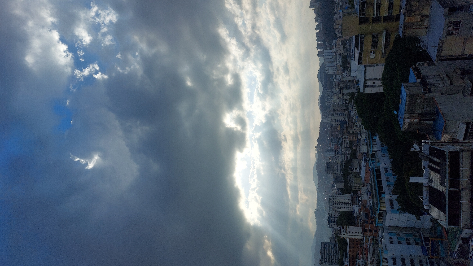 The image depicts an urban cityscape under a sky with dramatic cloud formations. The sky is divided between a dark, stormy area with heavy, blue-gray clouds on the left and a brighter, more luminous area on the right where sunlight streams through the clouds.
Below the sky, there is a dense urban environment, where there are buildings of varying heights and colors. These buildings create a densely packed urban landscape. The city appears to be Caracas, Venezuela.
No people are visible in the image.