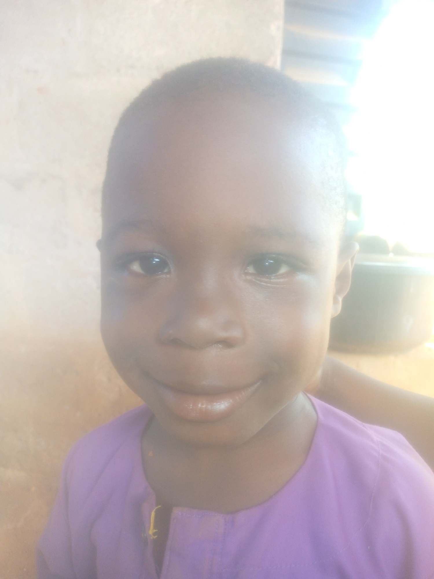 A close-up, brightly overexposed portrait features a young child with dark skin, looking directly at the camera with a wide, gentle smile. The child has short, dark hair and expressive dark eyes that reflect the intense light. They are wearing a purple garment, possibly a shirt or tunic, with a simple collar and a visible placket down the front. A small yellow embroidery or thread detail is visible near the neckline on the left side of the garment.
The background is heavily overexposed on the right side and upper half of the frame, indicating a strong light source, likely bright daylight. On the far left, a light-colored, textured surface, possibly a wall made of concrete or earth, is visible. Behind the child's right shoulder, a dark, indistinct object, possibly a container or pot, can be vaguely seen against the bright light. The child's smile conveys happiness or contentment. No text is visible in the image.