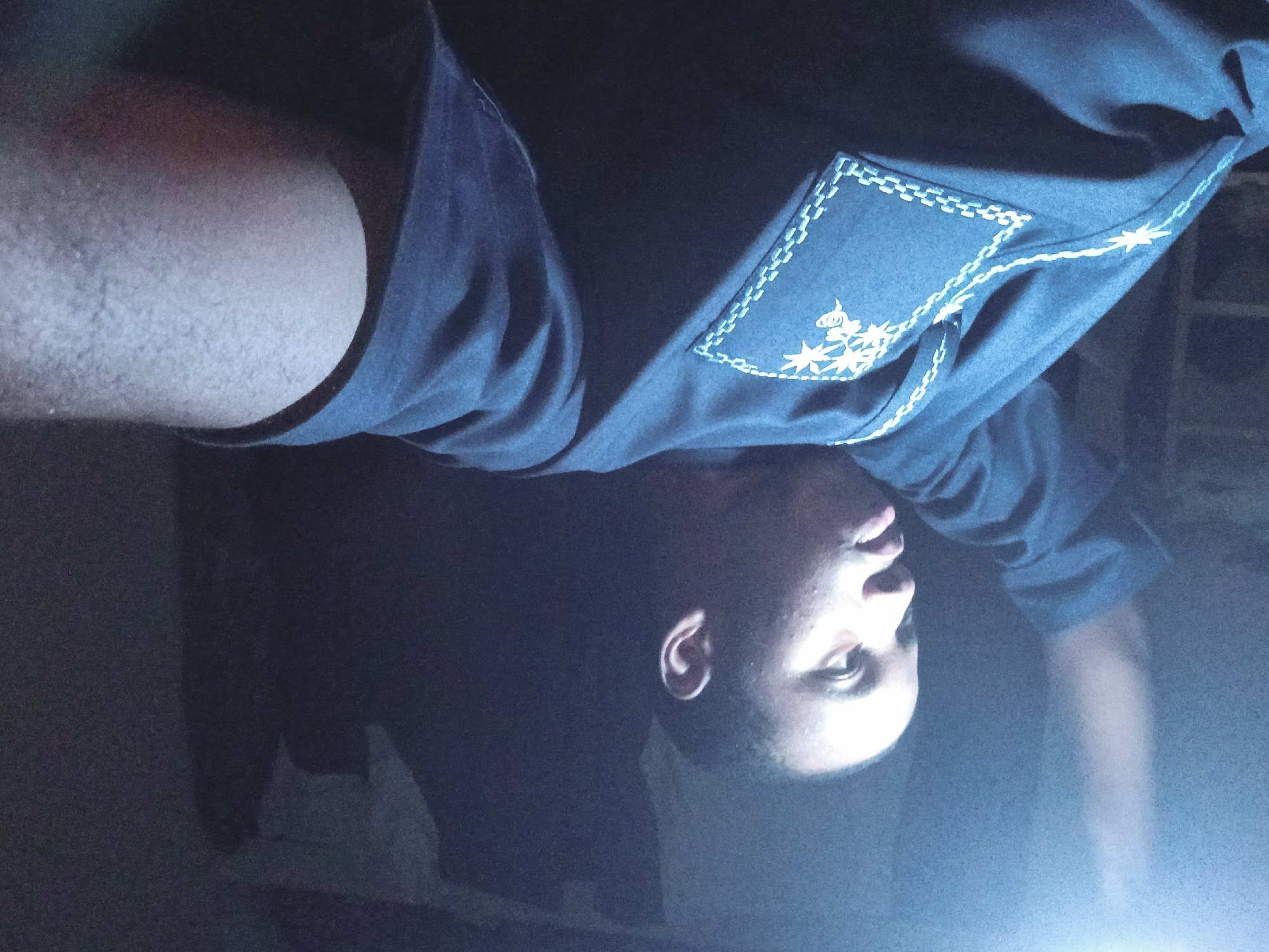 A person is photographed indoors. The individual, likely male, wears a dark-colored, long-sleeved shirt with light-colored embroidered detailing on the upper portion. Part of their arm and upper body are visible. The setting is poorly lit, with a dark background, and a blueish cast to the light. There is no discernible information to determine the location as Lagos, Nigeria.