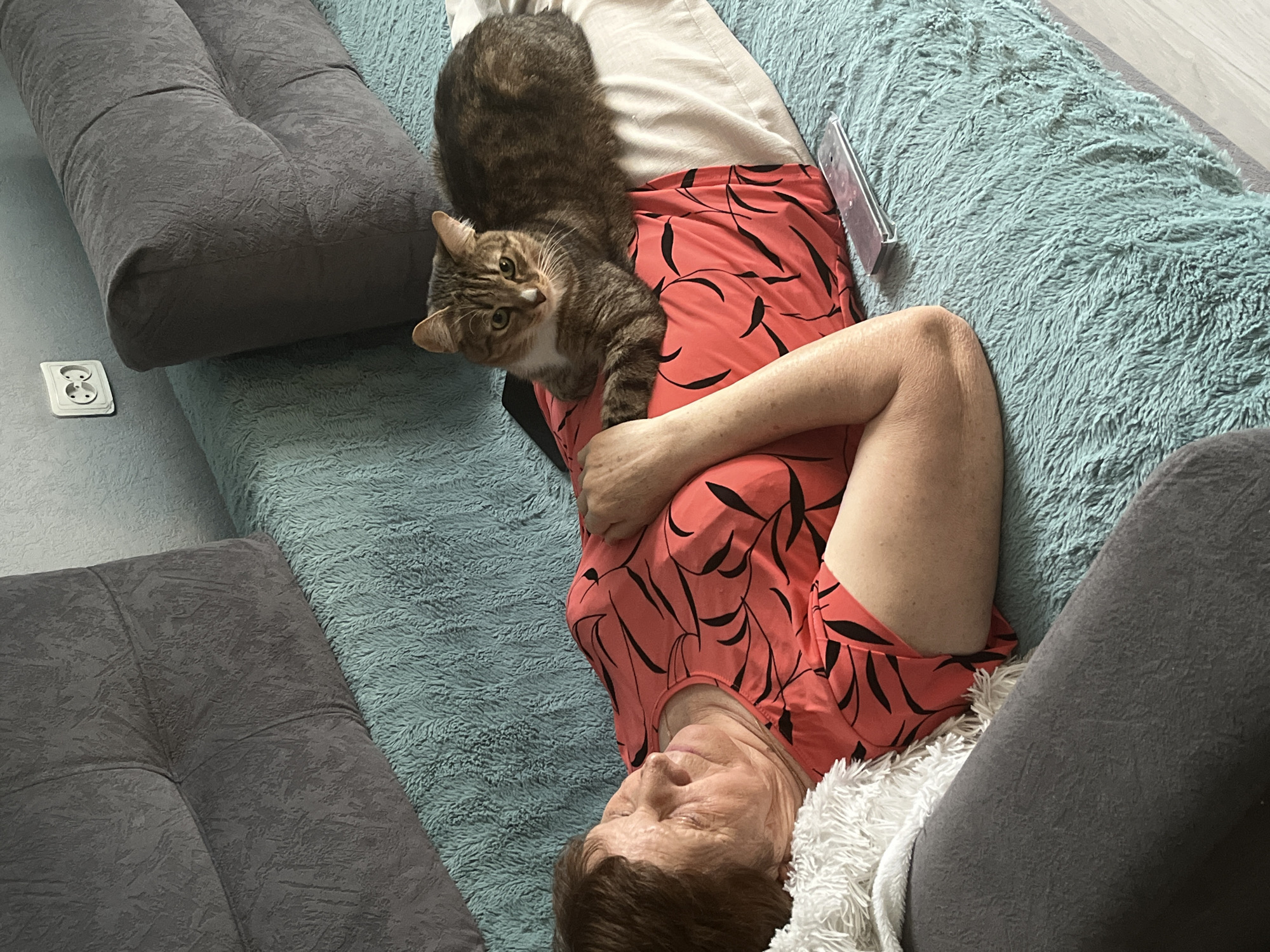 A woman is lying on her back on a turquoise shag rug, likely indoors. She is wearing a salmon-colored dress with black leaf-like patterns. A tabby cat is lying on her chest, looking towards the camera. Her left arm is across her torso and she appears to be resting. Part of a grey, tufted couch is visible on either side of the woman. A power outlet is visible on the floor next to the couch. A white blanket or rug is draped on her lower half near her grey pants. A smartphone is located above the woman. The location is not discernible from the image.
