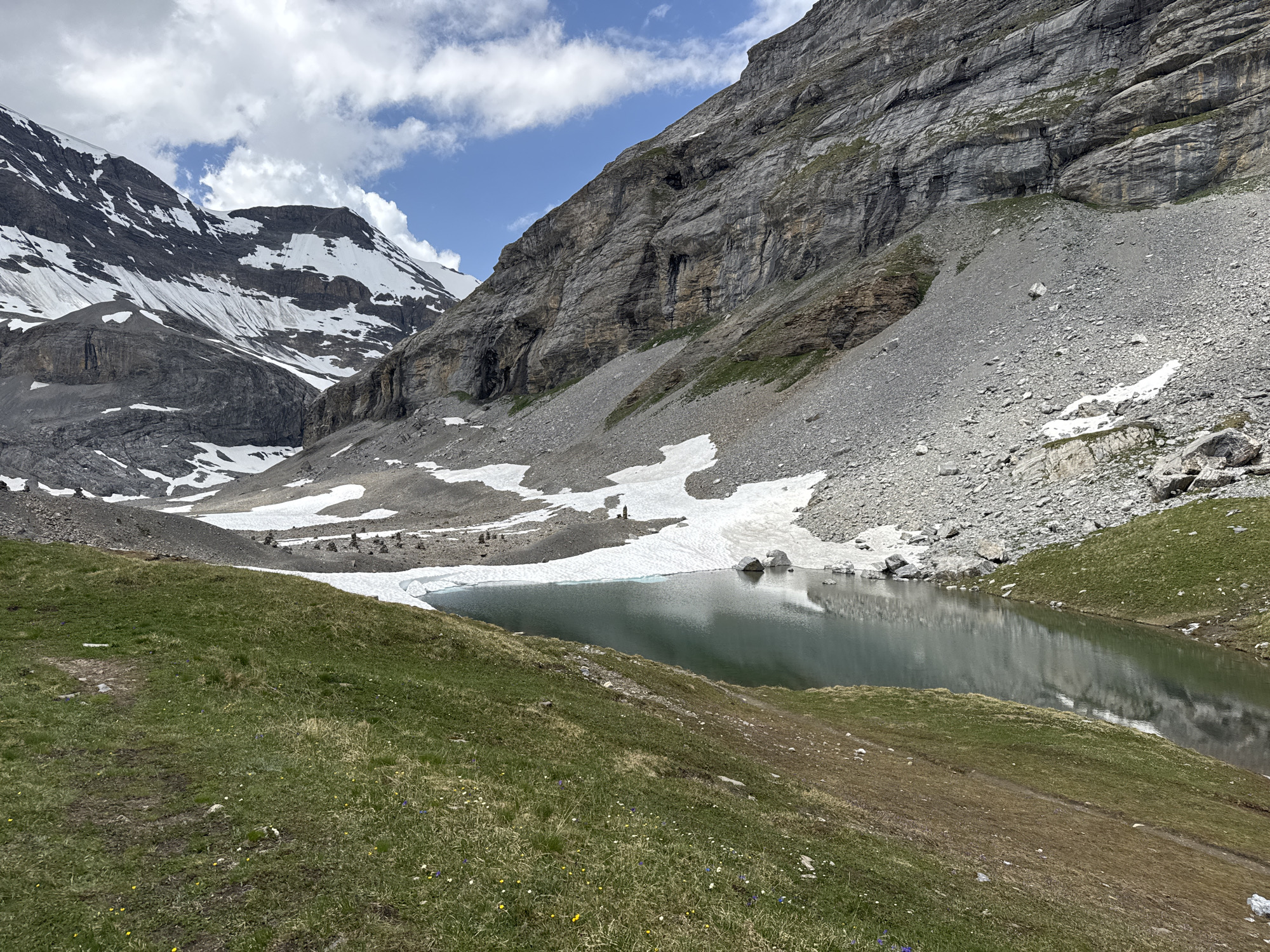 The image depicts a serene alpine landscape in the vicinity of Leukerbad, Switzerland. A dark, still lake occupies the lower right quadrant of the frame, its surface reflecting the overcast sky and the imposing, rocky mountains that dominate the background. Patches of snow cling to the slopes, suggesting a high-altitude environment. The foreground is covered in lush green grass, dotted with small wildflowers. A faint, winding path is visible on the grassy slope leading towards the lake. The overall atmosphere is one of rugged natural beauty and tranquility. There are no visible people or animals in the image.