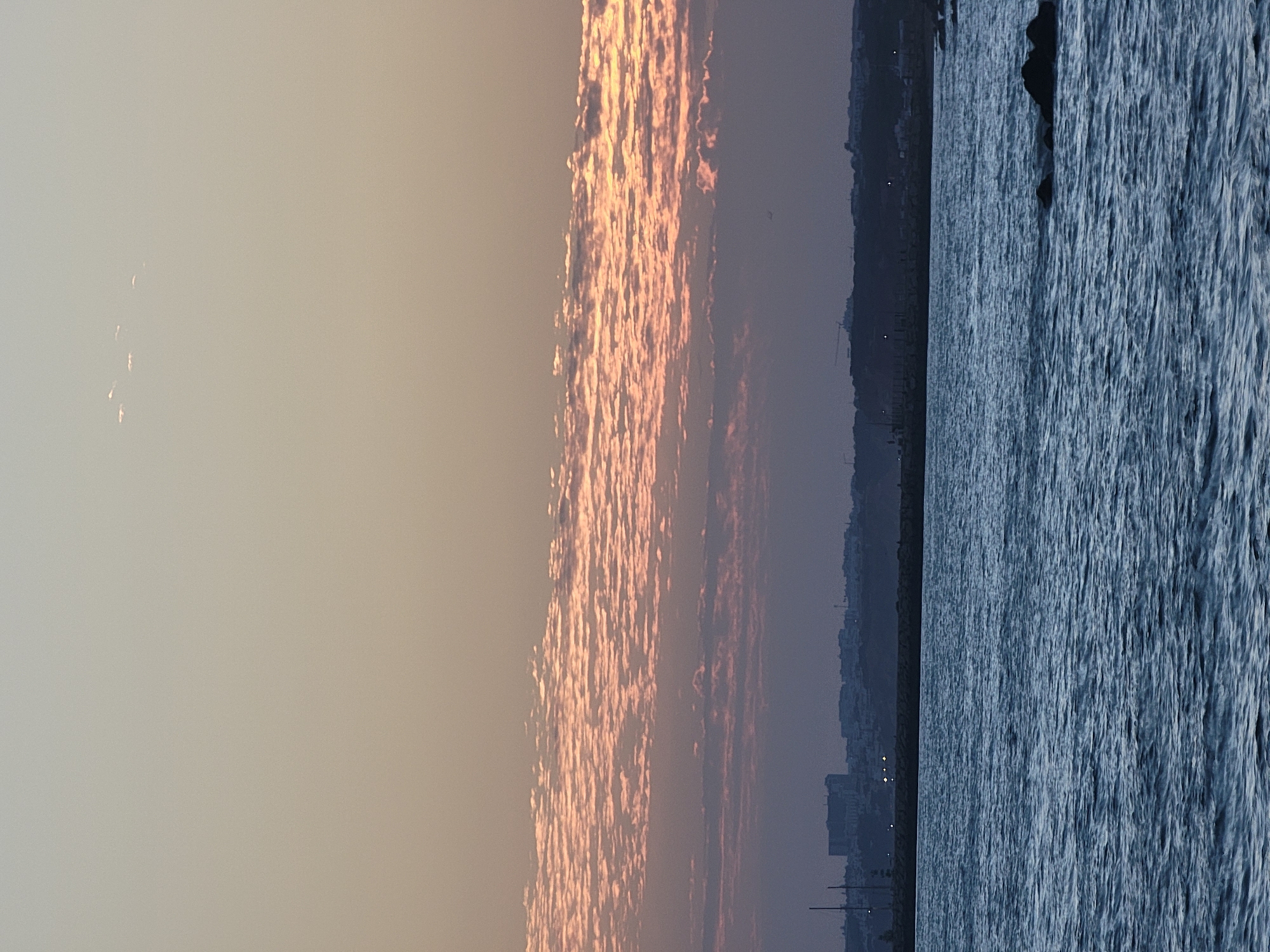 The image captures a serene seascape with a distant city skyline under a twilight sky. The foreground is dominated by a choppy, deep blue sea, with dark rocks emerging from the water on the right side of the frame. A concrete breakwater or quay runs horizontally across the middle ground, separating the sea from the city.
The city of Quarteira, Portugal, is visible in silhouette against the horizon. The skyline features buildings of varying heights, with some taller structures and what appear to be cranes, suggesting ongoing construction or industrial activity. Lights are visible along the waterfront, indicating an urban presence as dusk settles.
Above the city, the sky transitions through soft hues of pale yellow and peach, with bands of wispy clouds catching the last rays of the setting sun. These clouds are tinged with pink and orange, creating a textured, layered effect. Higher up, a few smaller, isolated clouds are scattered across the lighter sky. A small dark object, possibly a distant bird or aircraft, is faintly visible in the sky above the city. The overall atmosphere is one of tranquility and the transition from day to night.