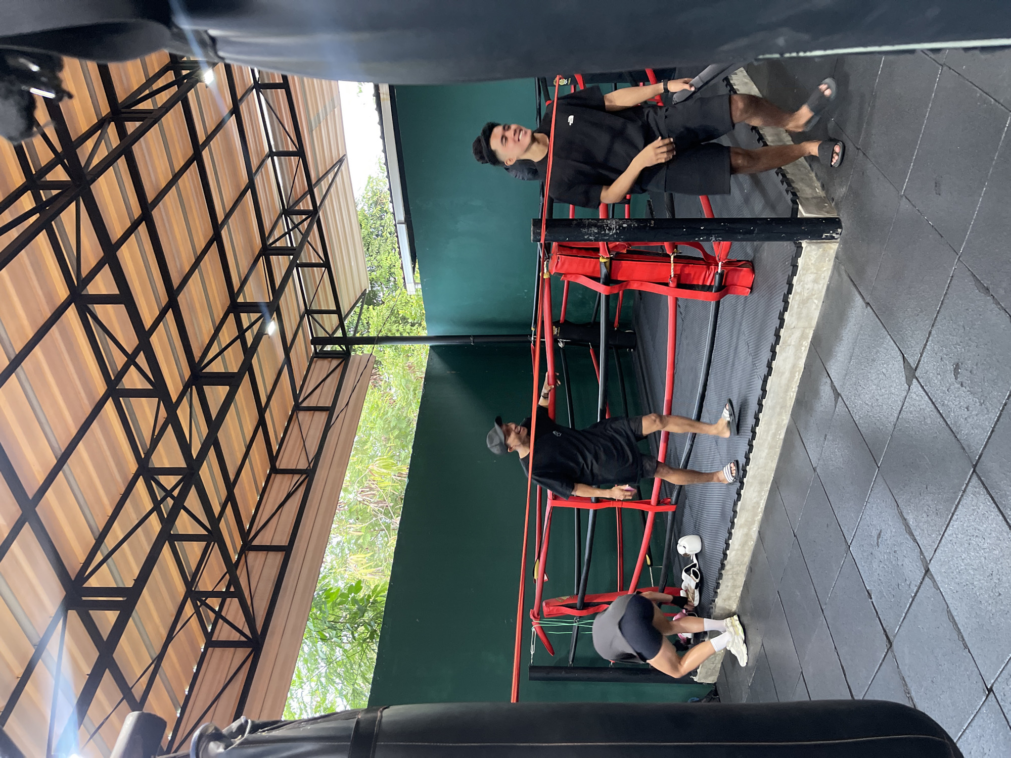 In the city of Kuta Selatan, Indonesia, within a boxing gym, three young men are present. One man is inside a boxing ring, leaning on the ropes, wearing a black t-shirt and black shorts, and smiling. Another man is also inside the boxing ring, wearing a black t-shirt and black shorts, with his head bowed as if looking for something on the floor. The third man is standing near the edge of the ring, wearing a black t-shirt and black shorts, holding a black punching bag. The boxing ring has red ropes and black padding. The gym floor is covered with black rubber mats. Above the ring, a roof structure made of black metal beams and translucent orange panels is visible, with some lights attached. Lush green foliage can be seen beyond the gym walls. Two large black punching bags are also visible in the foreground.