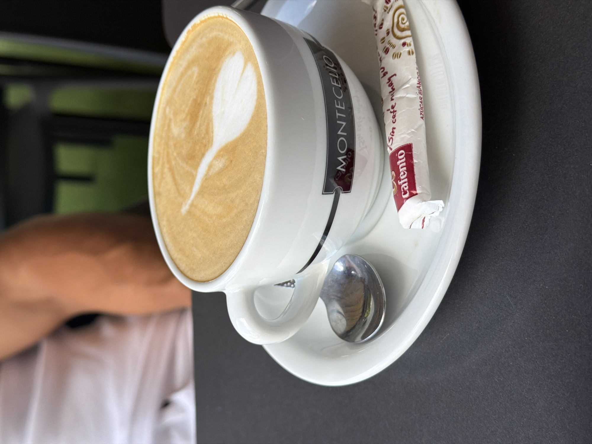 A white cup and saucer hold a latte with heart-shaped foam art and a packet of "Cafento" sugar. The cup features a black band with the white text "MONTECELIO". A shiny silver spoon rests on the saucer. The scene is set on a dark gray table, with a blurred background showing a person in a white shirt and an arm. The lighting suggests daytime, and the presence of a cafe setting implies an indoor or outdoor seating area. The overall mood is relaxed and inviting, characteristic of enjoying a coffee break. The sugar packet also contains Spanish text, including "Sin cafe, ni hoy", and "AZUCAR BLANCO".