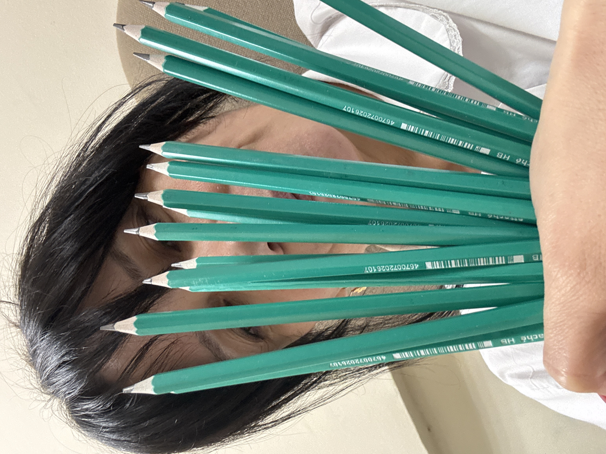 The image shows a person with dark hair partially obscured by a handful of green HB pencils. The pencils are held in front of their face, with the sharpened tips pointing towards the left. The pencils have a barcode and the text "4670072026107" printed on them. The person appears to be wearing a white shirt. It is impossible to determine if the image was taken in the city of Chaikovsky, Russia, or any other specific location.