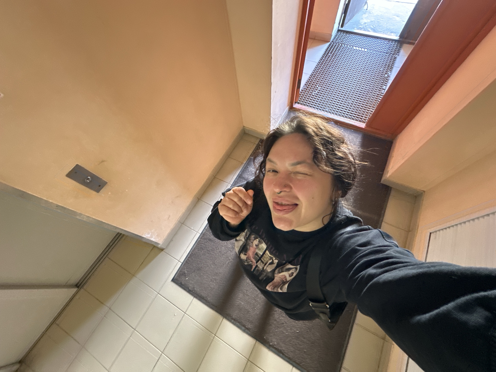 This image, captured in an indoor setting within the city of Chaikovsky, Russia, depicts a single individual from a low, upward-looking perspective. The person, positioned centrally in the frame, has long, wavy brown hair, squinted eyes, and an open mouth with their tongue partially extended. A nose ring is visible on their left nostril. They are wearing a black long-sleeved top or sweatshirt featuring a light-colored graphic print on the torso. A black strap, consistent with a bag strap, is visible across their left shoulder. The individual's right hand is partially visible in the upper right quadrant of the image, appearing clenched.
The environment includes a tiled floor composed of light beige square tiles with darker grout lines. A dark grey rectangular mat is situated on the tiles. The walls are painted in various tones: a lighter beige or off-white wall is visible on the left, while the upper and right portions of the image feature a peach or orange-toned wall. A metallic rectangular plate, secured by a single bolt or screw, is mounted on this peach-toned wall. To the far right, a reddish-brown framed opening is visible, containing a metal gridded panel. A light-colored baseboard runs along the bottom edge of this opening.