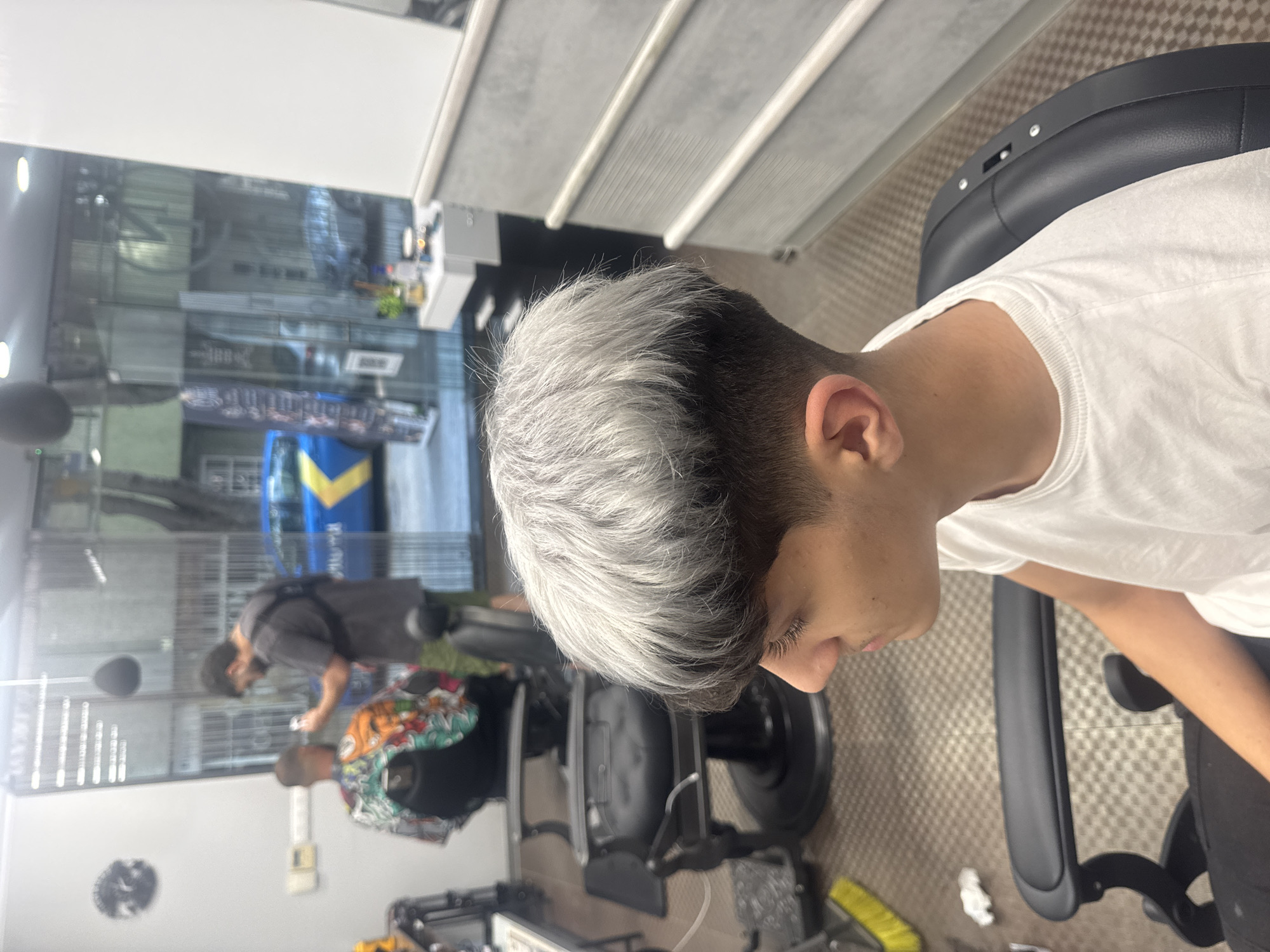 A young man with short, gray hair sits in a black barber chair inside a barbershop. He is wearing a white t-shirt. In the background, visible through the glass walls of the shop, there is a street scene with a blue and yellow vehicle and two people. One person is wearing a gray shirt, and the other is wearing a shirt with a colorful floral pattern. The barbershop setting includes mirrors and equipment. The precise location in the city of Varna, Bulgaria, is not identifiable from the image.
