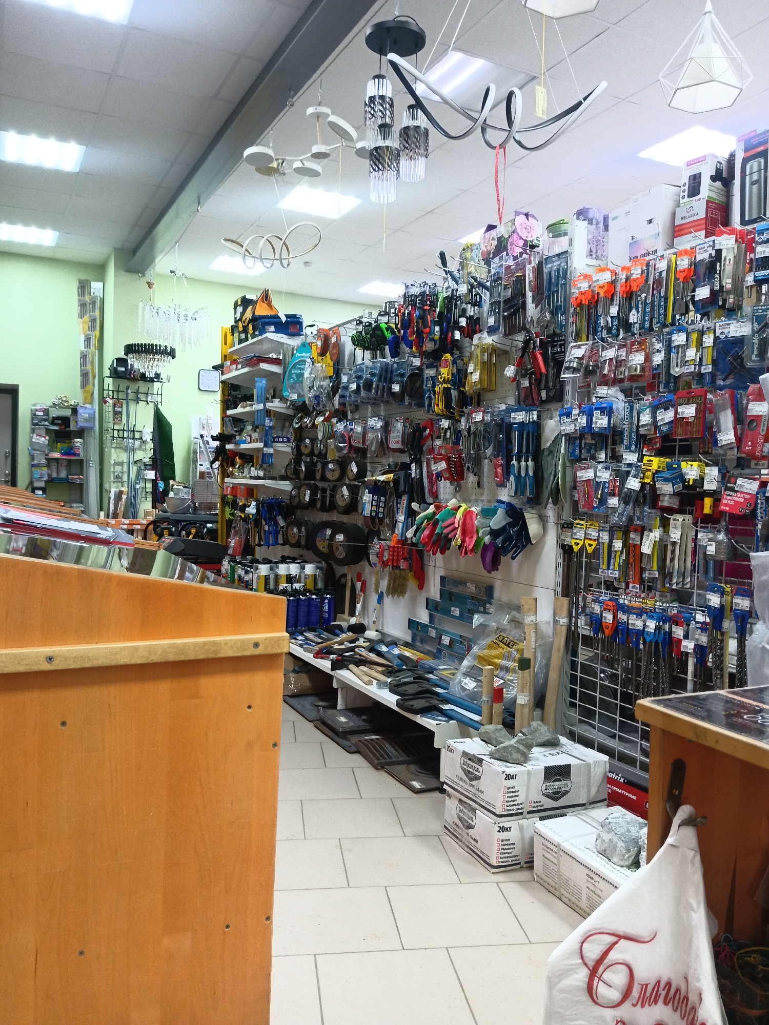 The image shows the interior of a store, possibly a hardware or home improvement store, located in Фоки, Russia. The scene is well-lit with fluorescent ceiling lights. The store is packed with a variety of merchandise displayed on shelves and hanging from wall-mounted grids. The items visible include tools such as screwdrivers, pliers, and drills, as well as gloves, measuring tapes, and other hardware supplies. There are also various household items and decorative light fixtures hanging from the ceiling.
In the foreground, there is a wooden counter with a metallic display. On the floor nearby are boxes labeled "20кг" with Russian text "КАМЕНЬ ДЛЯ ВАМИ," suggesting they contain stones for a sauna. A white bag with red Cyrillic script, seemingly "Благодар", is hanging from the counter. The floor is covered with light-colored tiles. The store appears to be fully stocked and ready for customers. No people are visible in the image.