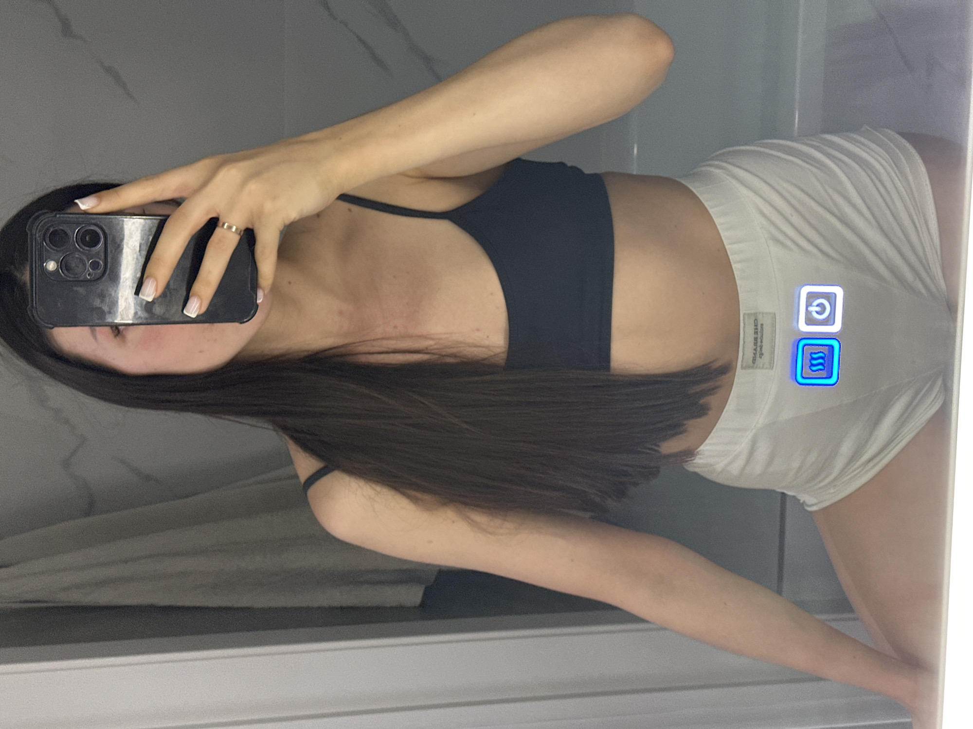 The image shows a person taking a mirror selfie in a bathroom. The person is wearing a dark bra and white shorts. The shorts have two illuminated buttons, one with a wavy line icon and the other with a power symbol. The background shows a marble tiled wall and a shower curtain. There is no indication of the location in Estachi, Russia.