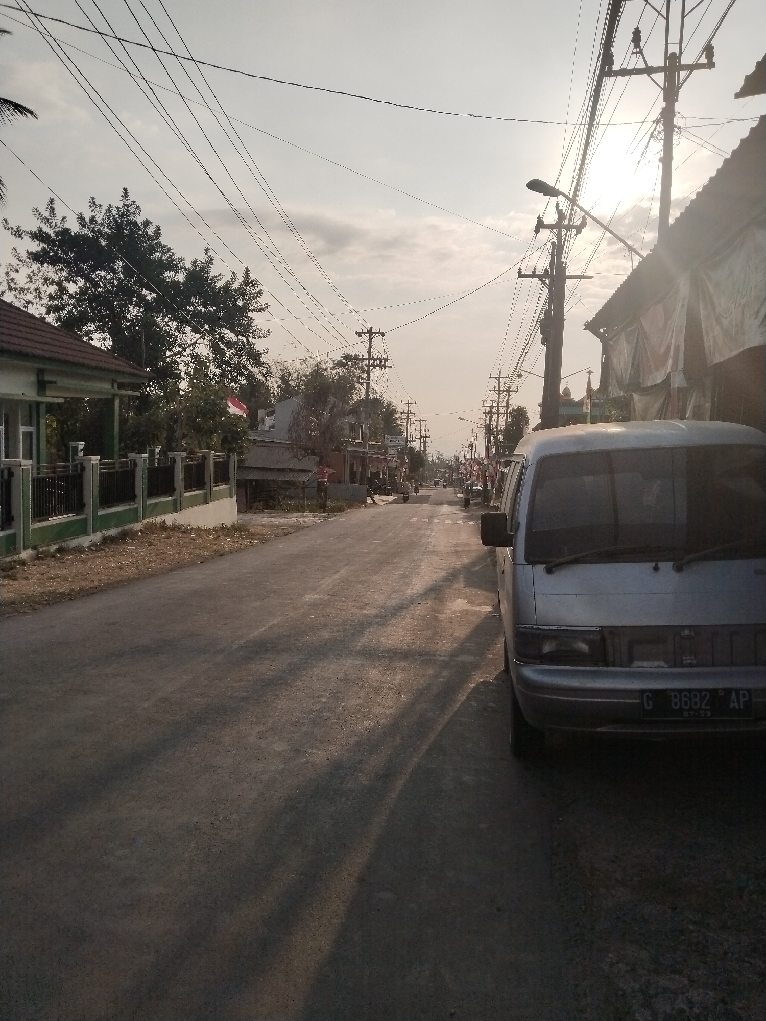 The image depicts a street scene in Harjowinangun, Indonesia, bathed in the soft light of either sunrise or sunset. A silver van, license plate "G 8682 AP", is parked on the right side of the paved road, its front facing towards the viewer. The road stretches into the distance, lined with buildings and trees. Utility poles with a complex network of wires dominate the upper portion of the frame, crossing the sky. On the left side of the road, a house with a green and white facade is visible behind a black metal fence. Further down the street, an Indonesian flag flies from a pole. The overall environment suggests a moderately populated urban or suburban area. The lighting casts long shadows across the road, indicating the low angle of the sun. There are no clear individuals depicted engaging in specific actions.