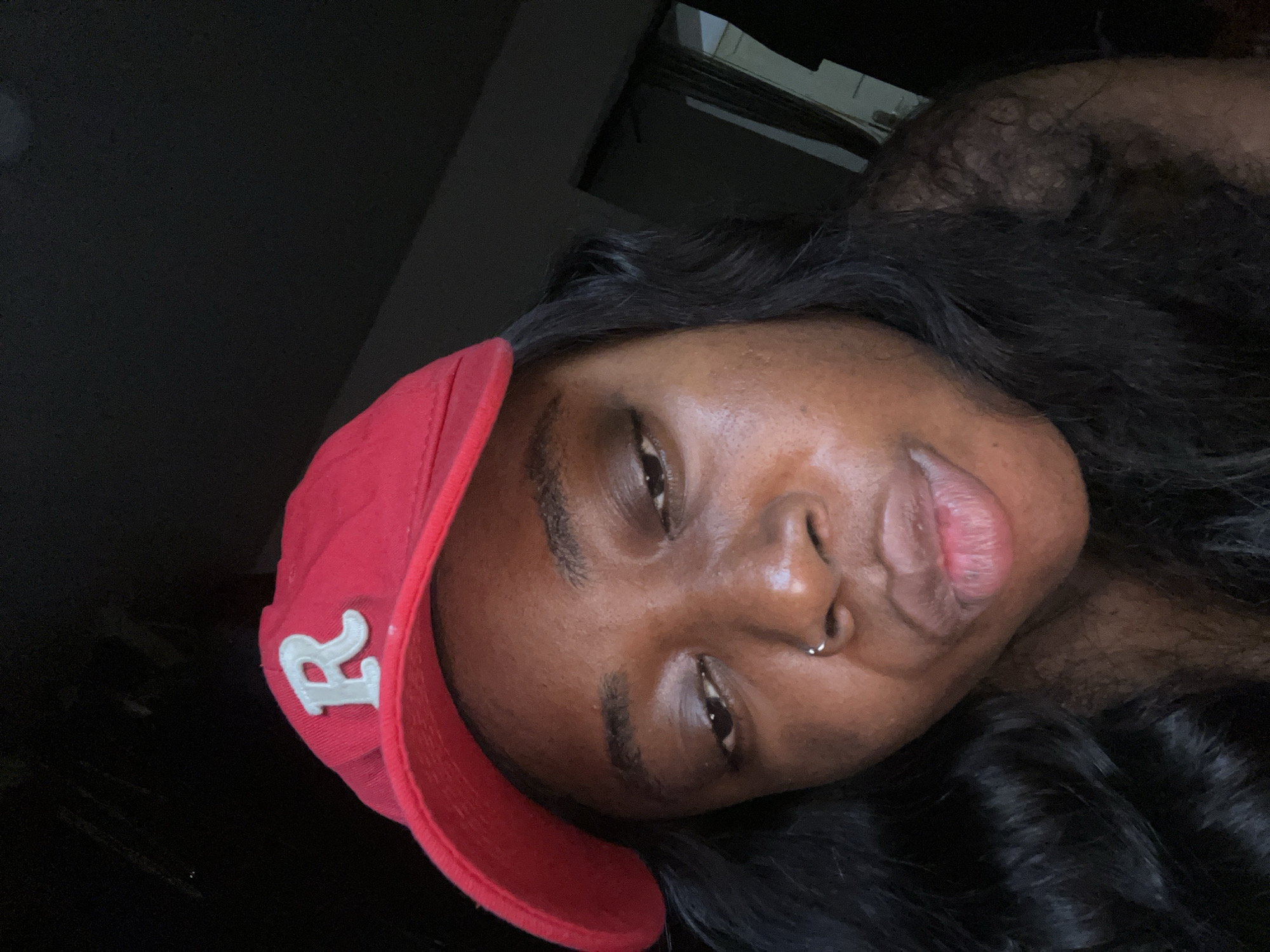 A close-up, selfie-style photograph shows a young Black woman wearing a red baseball cap with a white embroidered "R" on the front. She has dark, wavy hair, a nose ring, and her lips are slightly parted. The background is dark and out of focus, with a hint of a doorway visible on the right side of the image. The lighting appears to be artificial and low, casting shadows on her face. The overall mood is intimate and casual.