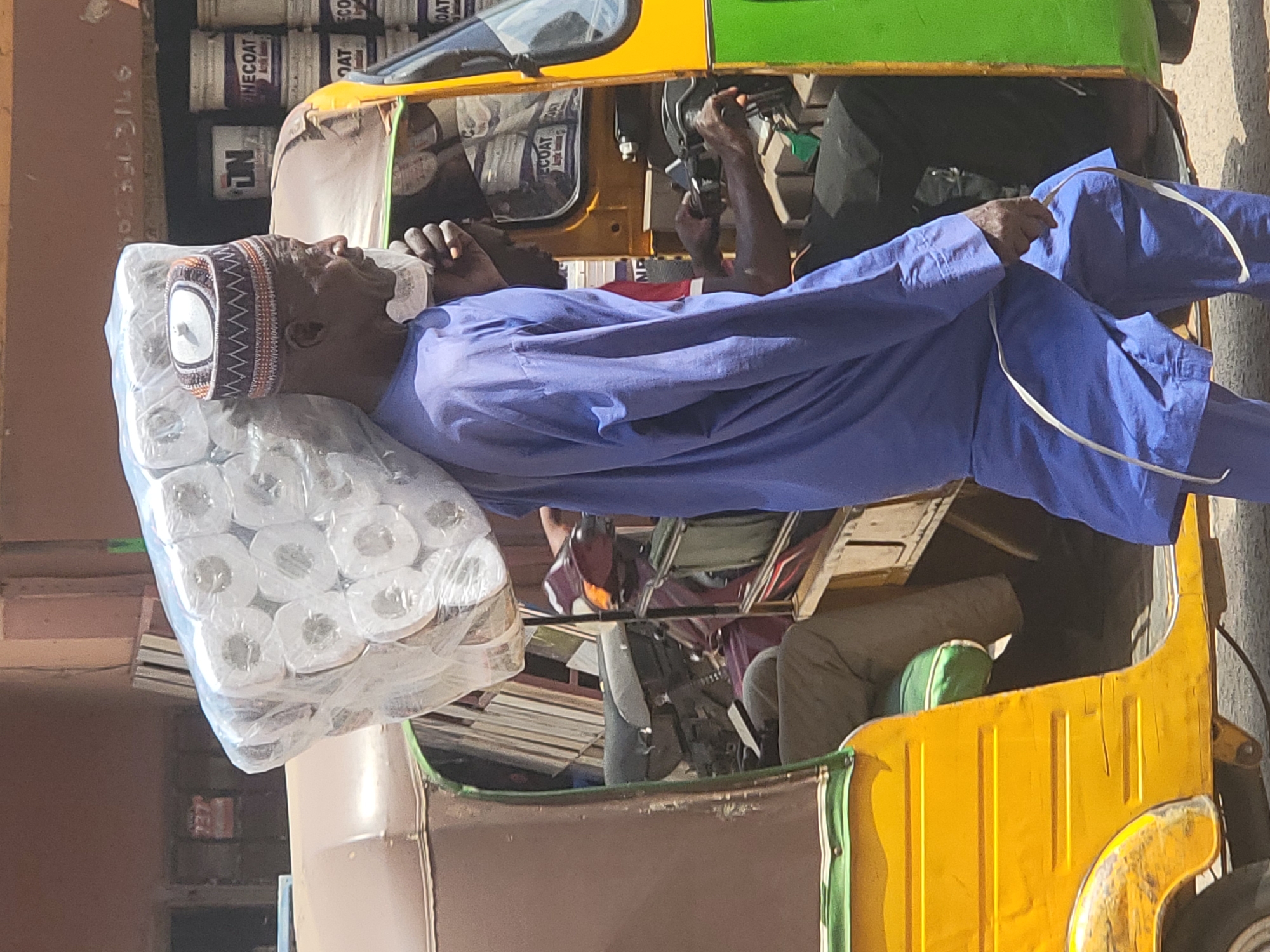 In Maiburiji, Nigeria, an elderly man with dark skin, wearing a patterned cap and blue robe, lies inside a yellow and brown three-wheeled vehicle. He is resting his head on a large, plastic-wrapped bundle of toilet paper. The driver is visible, holding the steering mechanism, with cans of paint stacked above the passenger compartment.