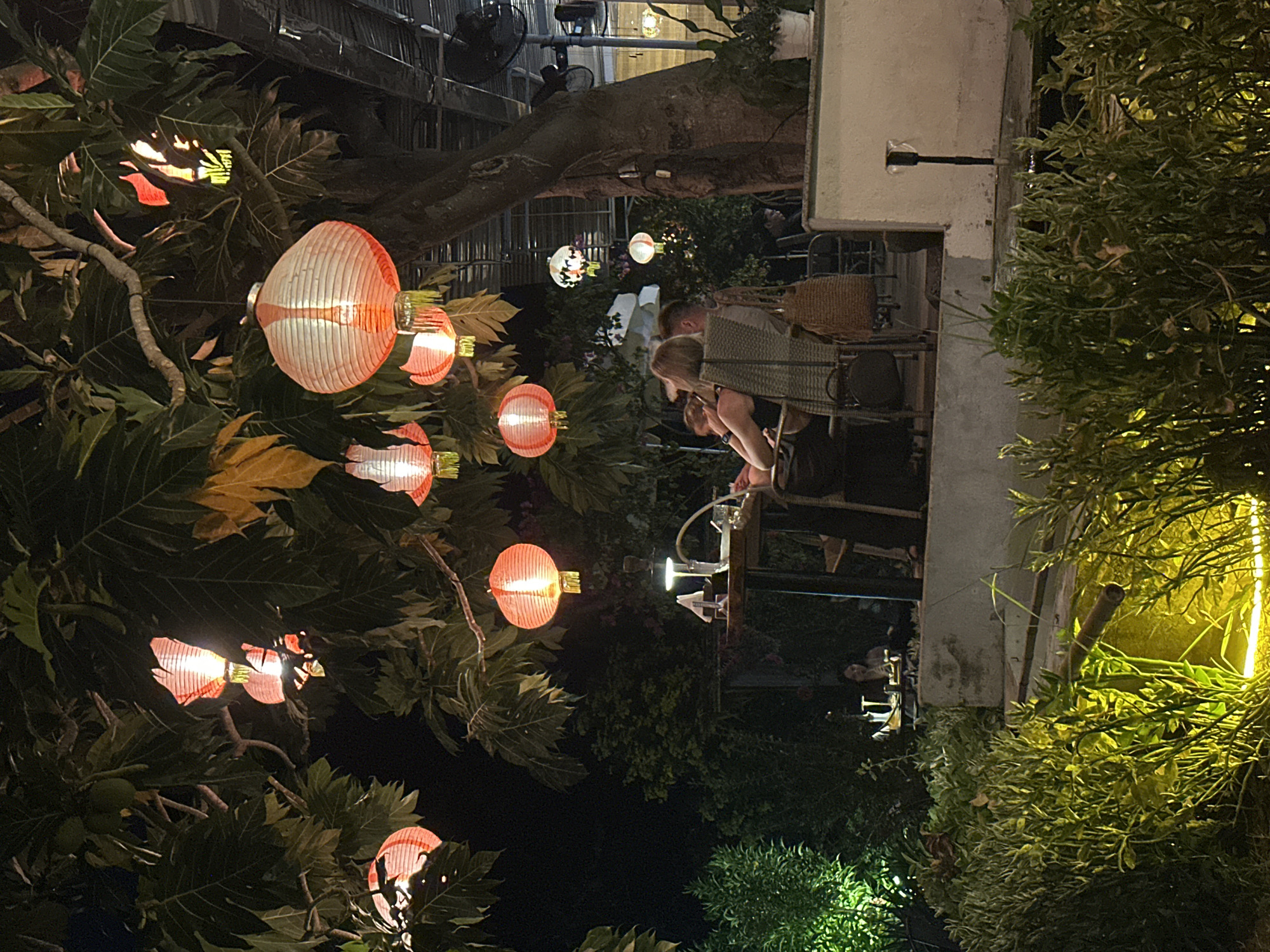 At night in Bac Nha Trang Ward, Vietnam, the scene captures an outdoor dining area illuminated by hanging lanterns. The lanterns are interspersed among lush foliage. People are seated at tables, with their activities partially visible, suggesting a restaurant or bar setting. Structural elements of a building are visible, integrated with the greenery.