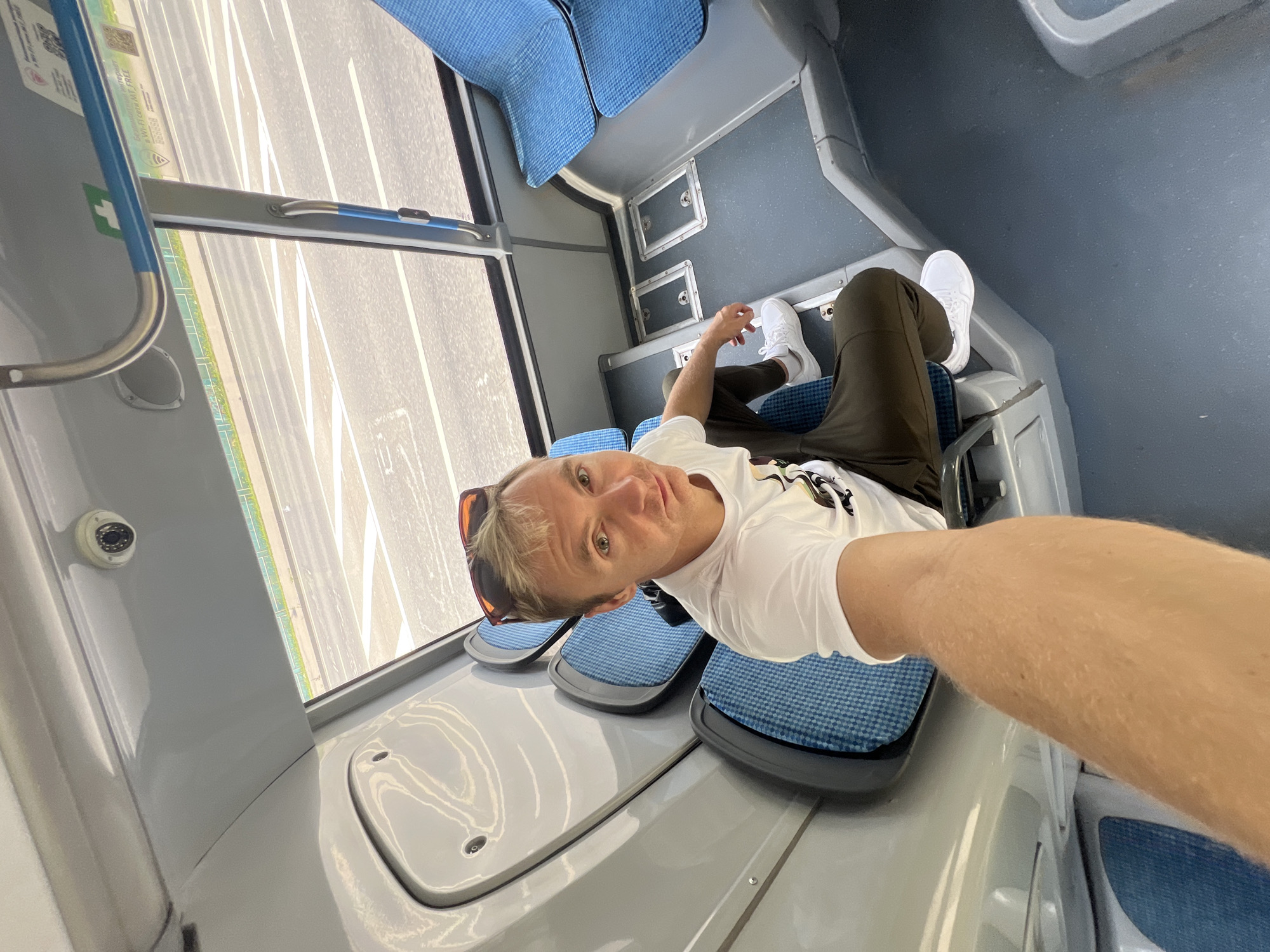 In the Vnukovo, Russia, a man with blond hair and sunglasses perched on his head sits in the passenger area of a bus. He is wearing a white t-shirt and dark pants, with white sneakers on his feet. The man is looking directly at the camera, and appears to be taking a selfie. The bus interior features blue seats, grey paneling, and a security camera mounted on the ceiling. Through the bus windows, a road with white markings and some greenery are visible, suggesting an urban or suburban setting.