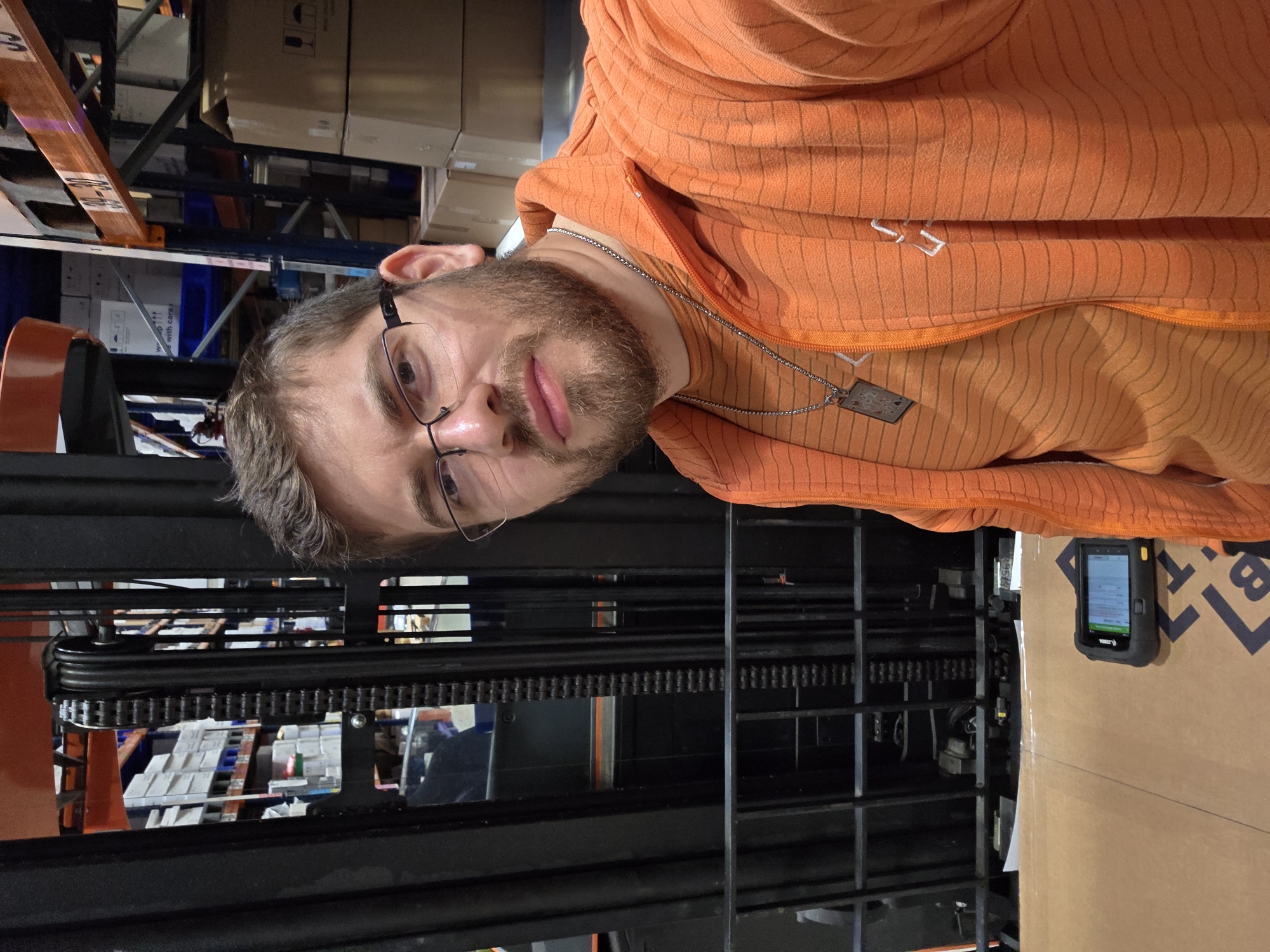 A man with a beard and glasses wearing an orange striped hoodie is positioned in front of a forklift mast and chain mechanism. In the background, shelves are visible, stocked with boxes. A handheld scanning device is affixed to a large cardboard box to the right of the forklift mast. The image appears to be taken indoors, likely in a warehouse or storage facility, but there's no direct indication it's specifically in Plovdiv, Bulgaria.