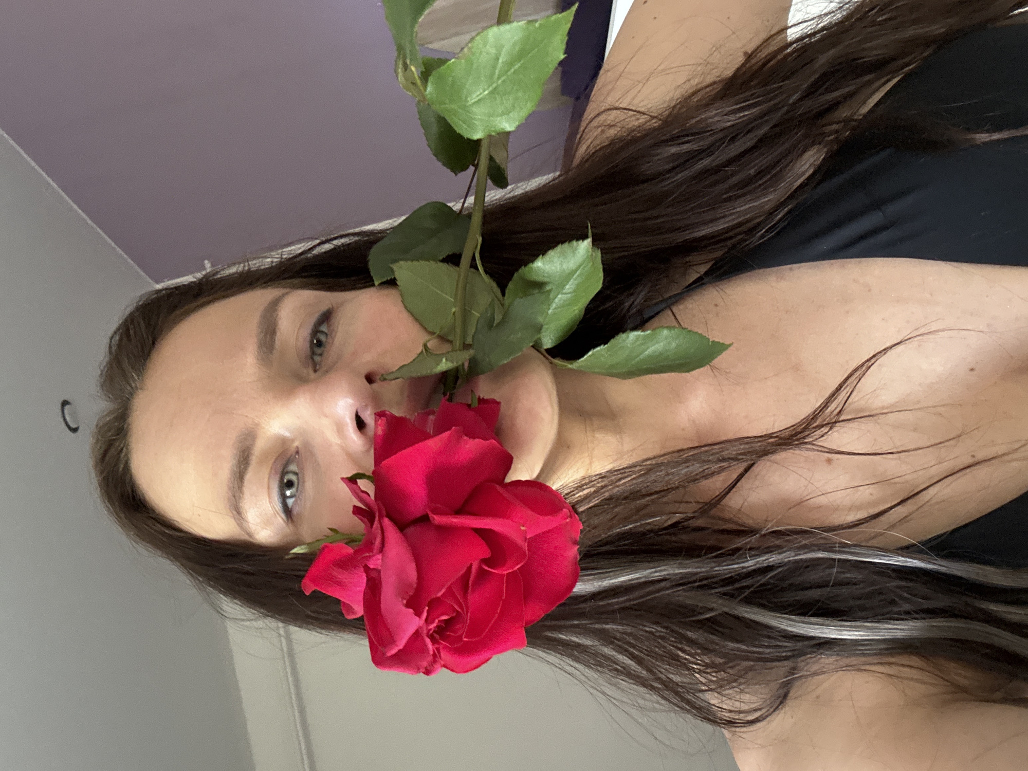 A person is holding a red rose close to their face. The background is out of focus but shows a wall. The person has long brown hair with a few silver streaks. It is impossible to ascertain the location in the city of Rusalevka, Russia from this image.