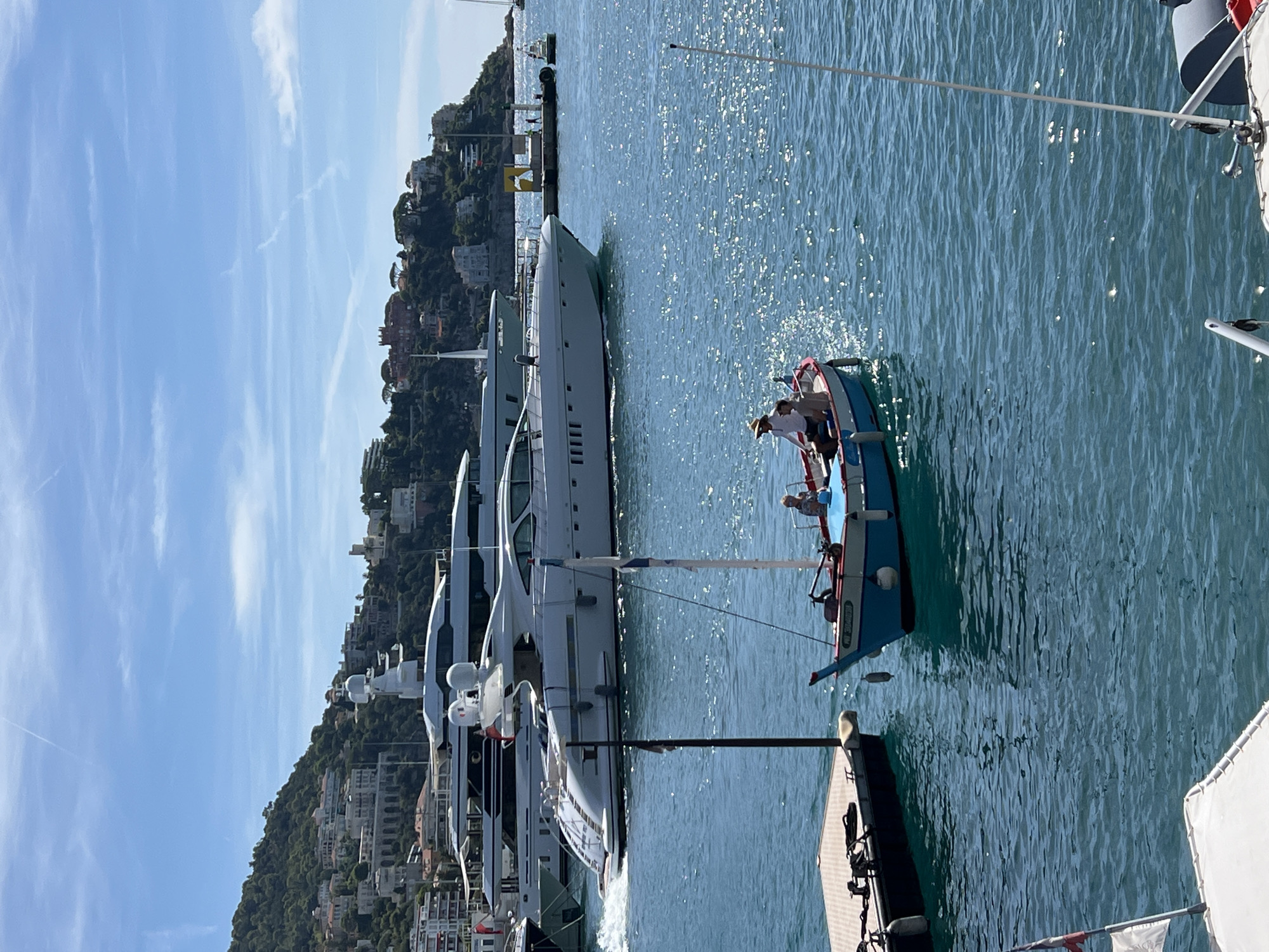 The image depicts a sunny day at the port of Nice, France. The foreground shows a small, blue and red boat with two people aboard, one wearing a straw hat. In the background, several large, white yachts are docked, with a hillside covered in buildings rising behind them. The water is a vibrant blue, reflecting the sunlight. A dock is visible to the left of the smaller boat. The sky is clear with a few wispy clouds.