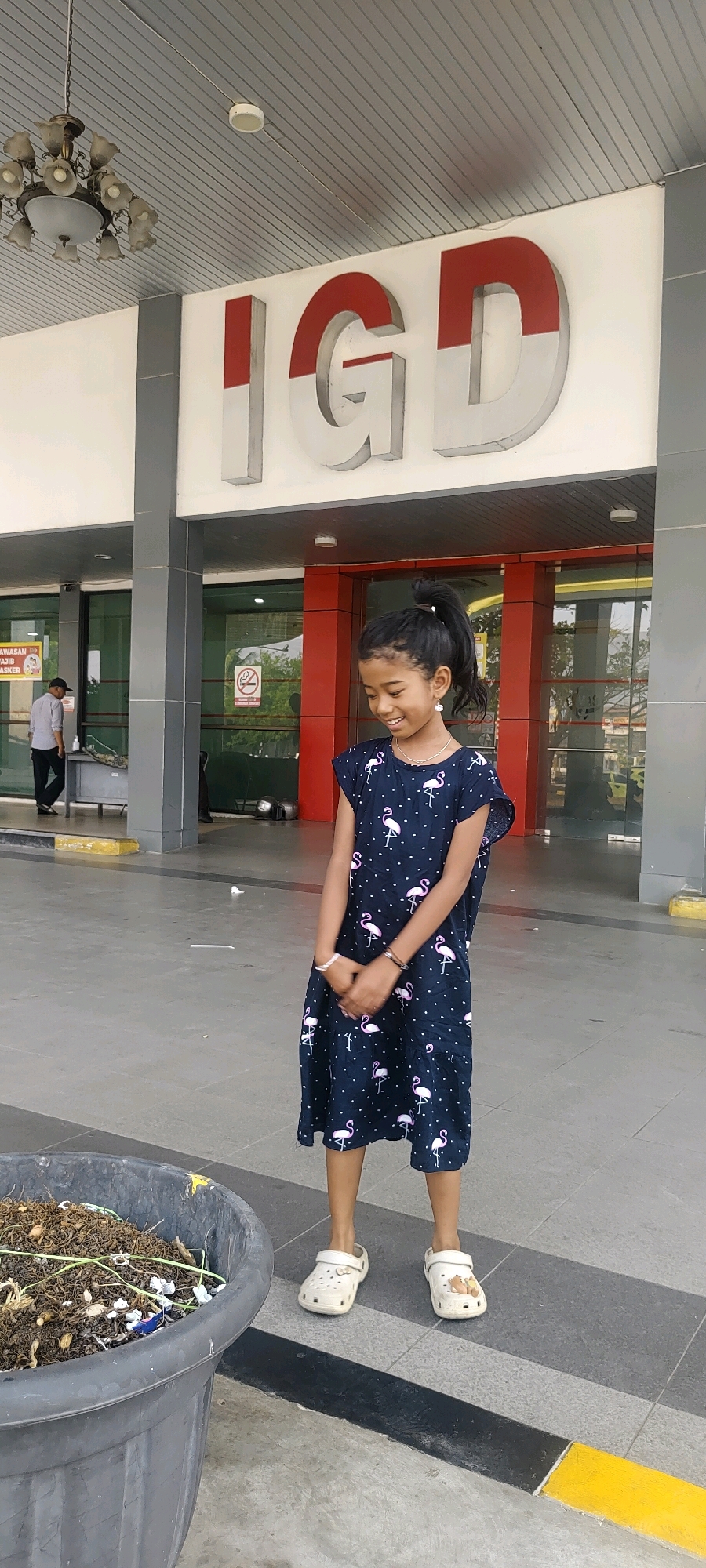 A young girl with dark hair pulled into a ponytail stands in front of a building with large, red and white letters spelling "IGD." She is wearing a dark blue dress with white polka dots and pink flamingoes. Her hands are clasped in front of her, and she is looking down with a smile. She wears white slip-on shoes. To her left is a large, dark grey planter filled with soil and dried plant matter. Behind her, the building has large glass windows and red accents around the entrance. A man in a grey shirt and black pants is visible through the window to the left. The ground is paved with grey tiles and has black and yellow lines indicating a pathway. The lighting suggests it is daytime.