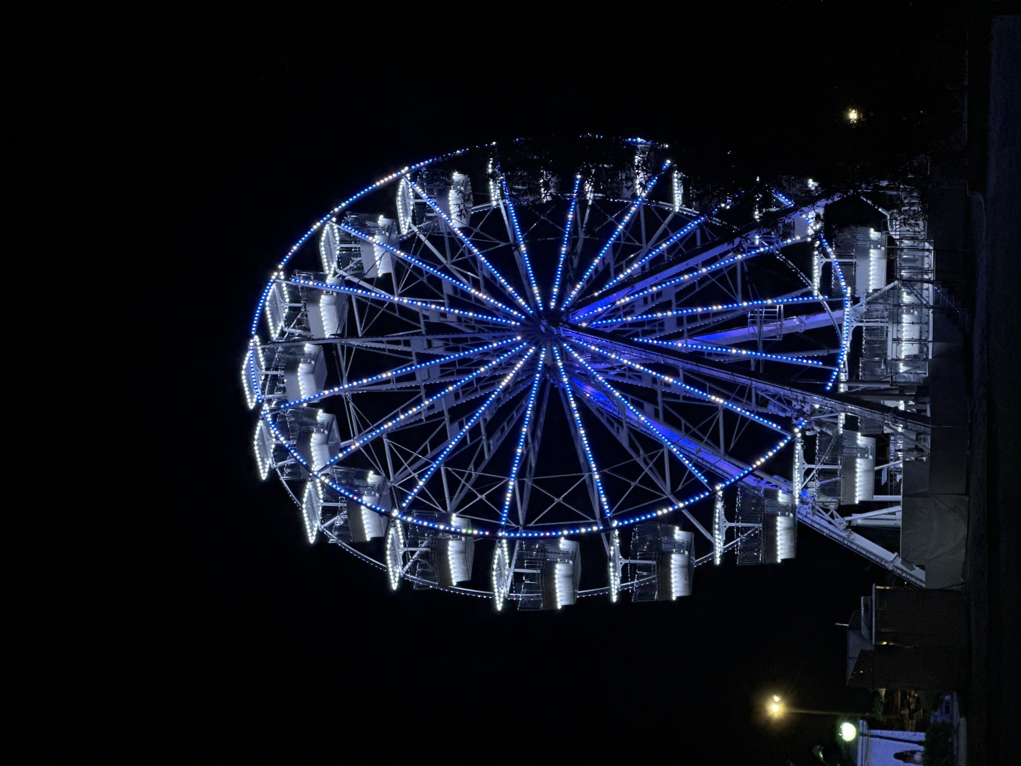 A Ferris wheel is lit with blue and white lights at night, in an outdoor setting. The Ferris wheel is the dominant subject, with its circular structure and radiating spokes illuminated by numerous small lights. The gondolas are visible, suspended from the wheel, also lit from within. In the foreground, there are some dark structures and a street light is visible to the left of the Ferris wheel. The background is dark, indicating it is nighttime. The scene suggests a fairground or amusement park. The image is taken from a low angle, looking up at the Ferris wheel.