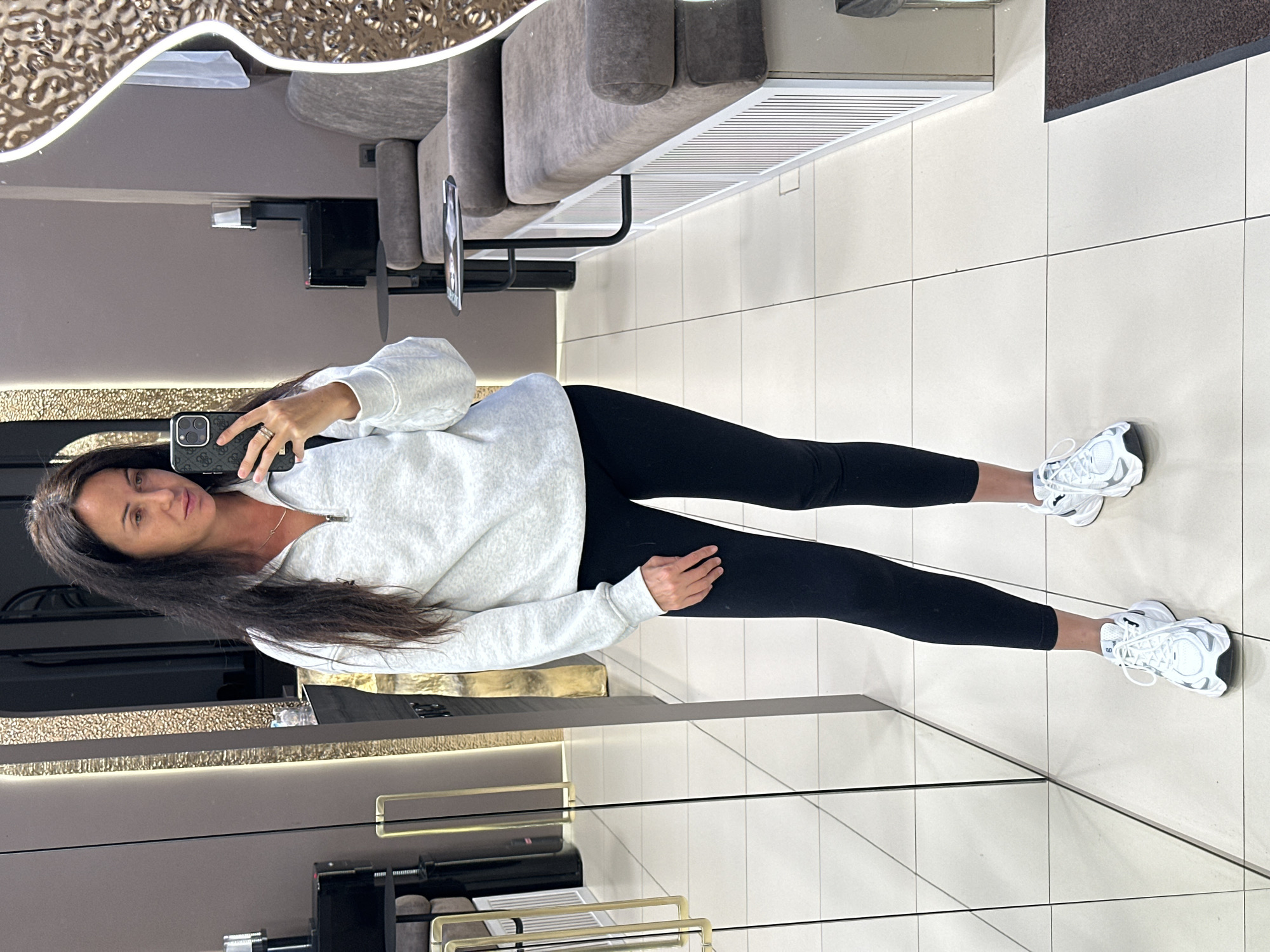 A woman with long dark hair is taking a selfie in a mirror. She is wearing a light gray quarter-zip sweatshirt, black leggings, and white athletic shoes. The background appears to be an indoor commercial space, possibly a salon or boutique, with mirrored walls, seating areas, and decorative elements. There are no visible indicators to suggest this location is in Irkutsk, Russia.
