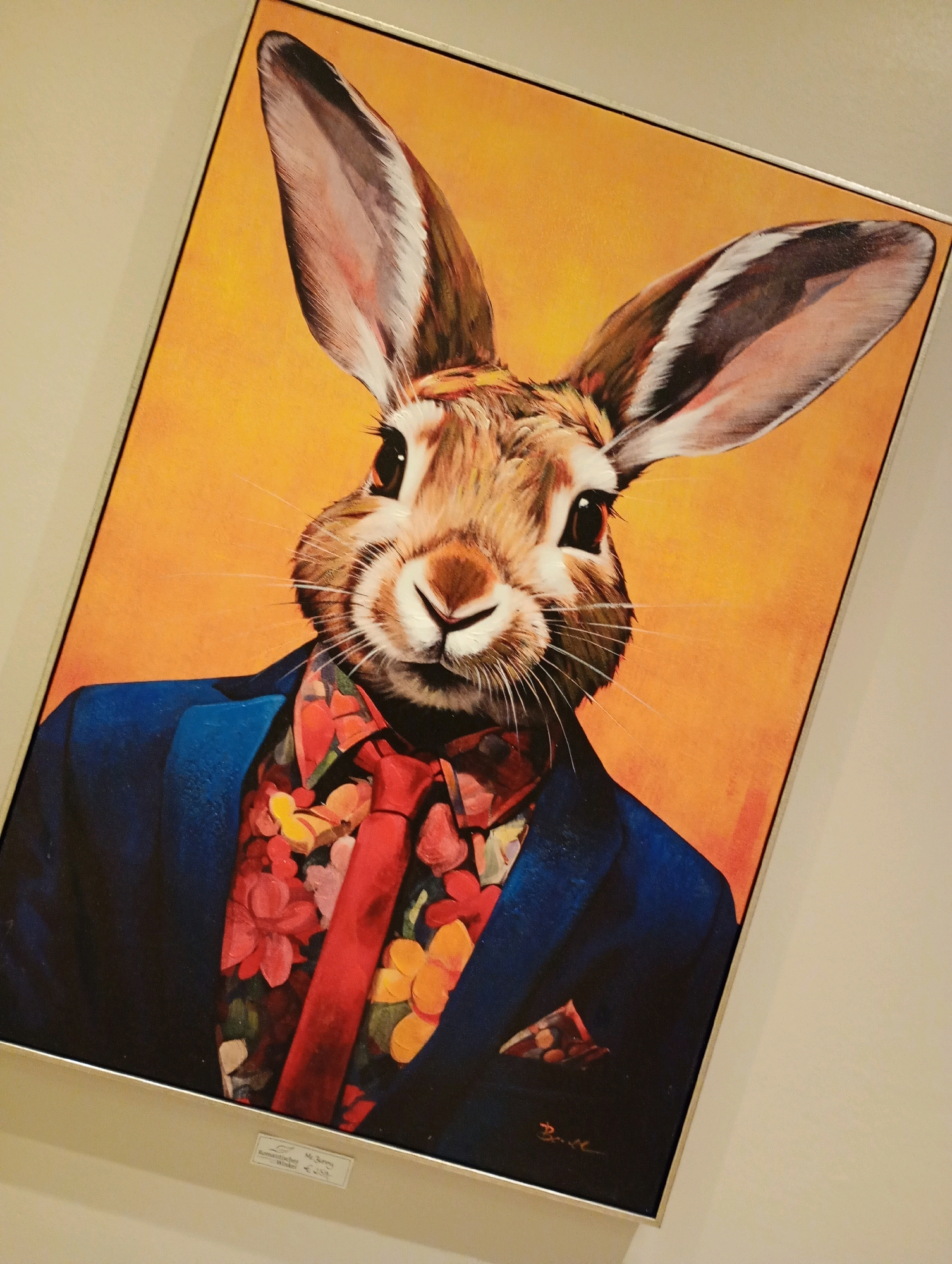 A framed painting of a rabbit wearing a suit, floral shirt, and red tie hangs on a light-colored wall. The rabbit has large ears and is looking directly at the viewer with dark eyes. The background of the painting is a bright orange color. A small label is affixed to the bottom of the frame, partially visible, indicating "Mr. Bunny" and a price of "€ 25,-". The painting is located in the city of Bad Sachsa, Germany.