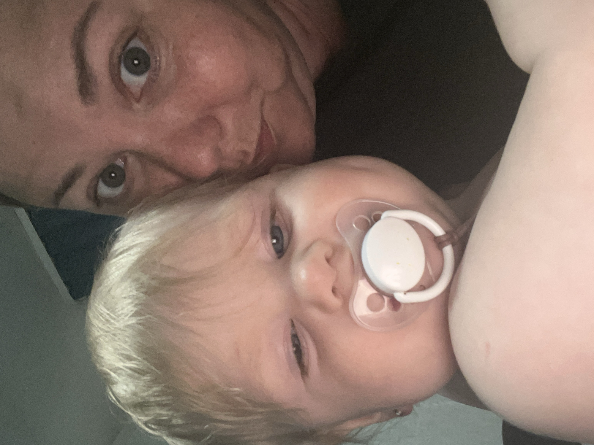 The image shows a close-up selfie of an adult and a baby. The adult, who appears to be a woman, is in the upper right corner of the frame, looking directly at the camera with wide eyes. The baby is in the foreground, taking up the left and center portions of the image. The baby has blonde hair and a pacifier in its mouth. The background is dark and indistinct, suggesting an indoor setting at night. The image quality is somewhat grainy, and the lighting is dim. No specific location in the city of Фоки, Russia, can be identified from the visual information provided.