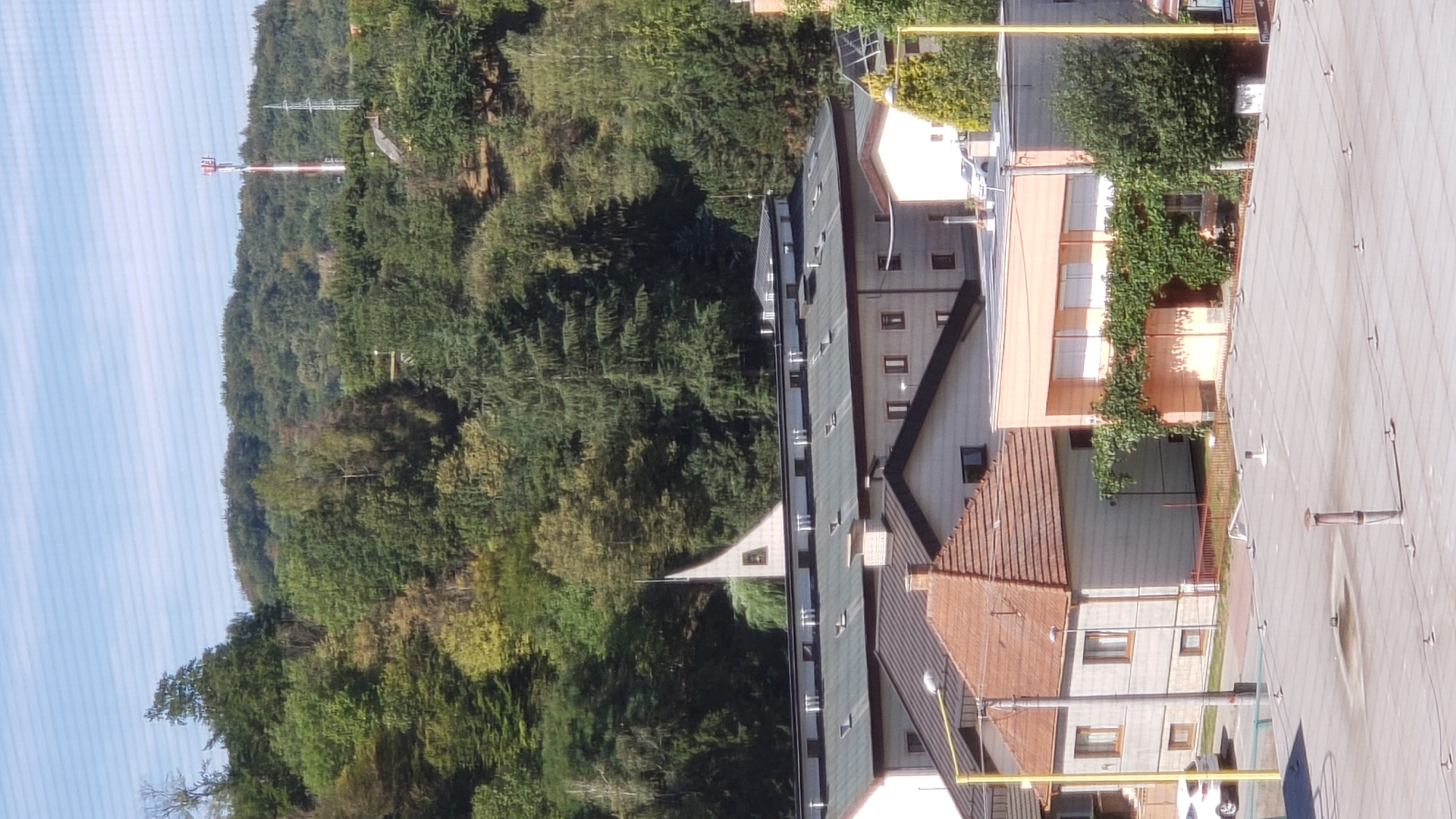 The image shows a view from an elevated position, looking towards a wooded hillside in the distance. In the foreground, there is a flat, grey rooftop with several ventilation pipes and electrical fixtures. Several buildings are visible in the mid-ground. To the left, a house with a brown tiled, steeply pitched roof and light-colored siding is visible, with a yellow pole and street light in front of it. Next to it is a longer, light-colored building with a flat roof, featuring several evenly spaced windows. Further to the right, a building with an orange-colored facade is adorned with climbing green vines. This building also has a flat roof with white-shuttered windows. A taller, grey building is situated to the right of the orange building. In the background, a densely wooded hillside dominates the scene. A red and white communication tower and a metal pylon are visible on the ridge of the hill. A pointed, white spire, possibly belonging to a church or similar structure, rises from behind a long, dark-roofed building in the mid-ground, partially obscured by trees. The sky is clear and blue with faint horizontal lines. The scene appears to be a residential or commercial area at the edge of a town, likely Prešov, Slovakia, given the presence of distinct architectural styles and the communication tower commonly found in urban landscapes. There are no people visible in the image.