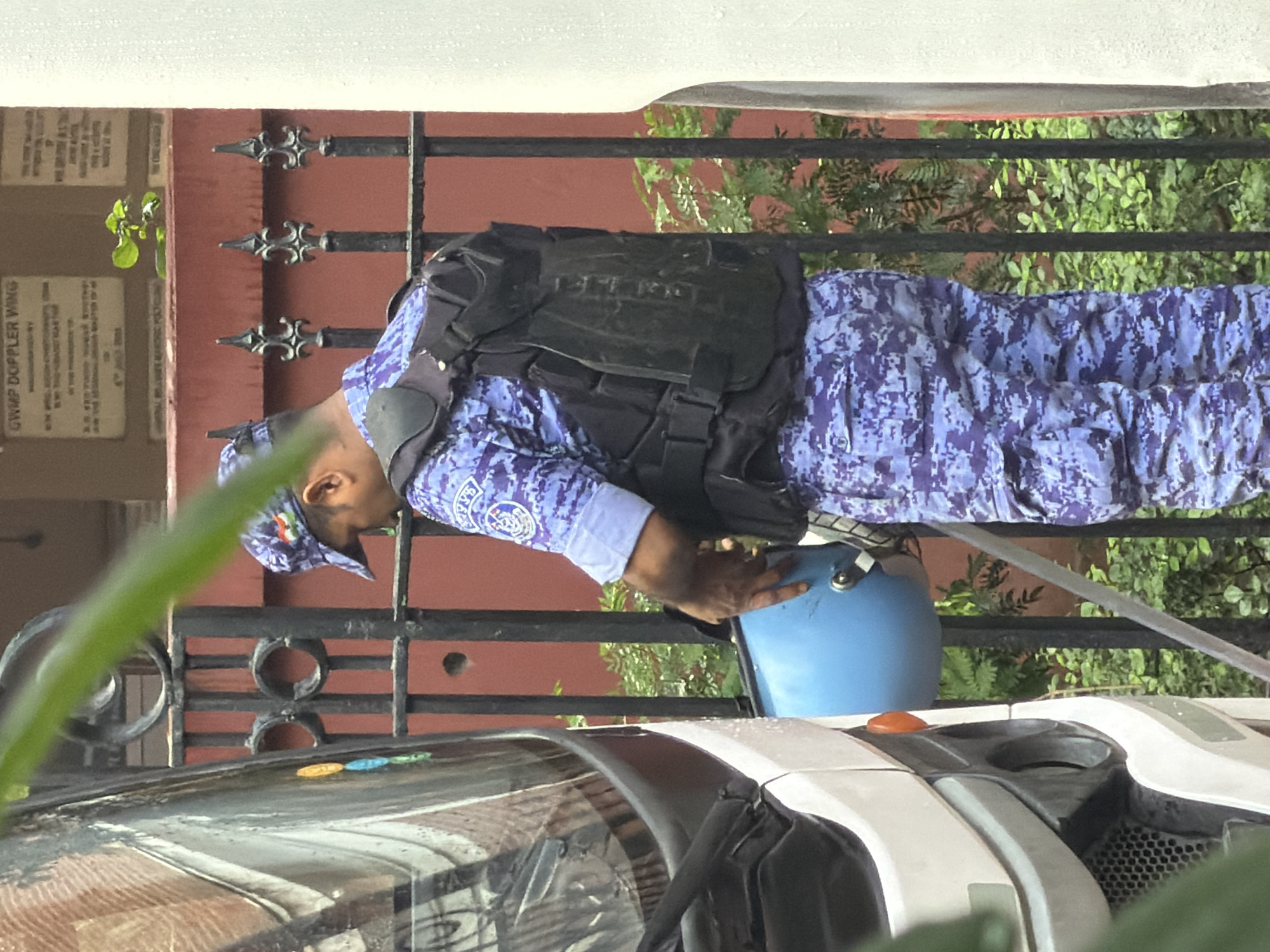 A person in a blue camouflage uniform with a black vest and helmet is standing next to a white vehicle. The person is holding a light blue helmet in their right hand and their head is bowed. The uniform has a patch on the sleeve. The person is standing behind a black metal fence. In the background, there is a red wall with some plaques on it and green foliage. There are raindrops on the right side of the image, suggesting it might be raining. The scene appears to be in an urban environment, possibly New Delhi, India, given the presence of the Indian flag insignia on the cap.
