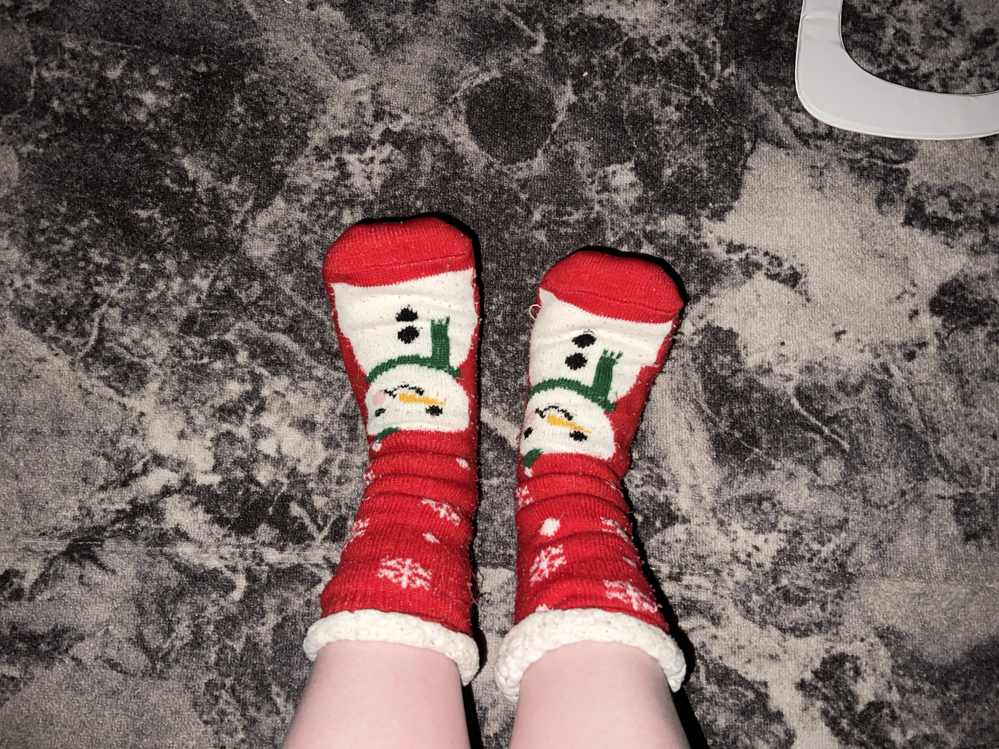 The image shows a pair of legs wearing red and white snowman-themed socks on a gray and black patterned rug. The socks have a white fluffy trim at the top, a red cuff, and a snowman design. The background is a dark gray and black rug with a mottled pattern. A white object is visible in the upper right corner, partially cropped. The legs are pale. The location cannot be inferred from the image.